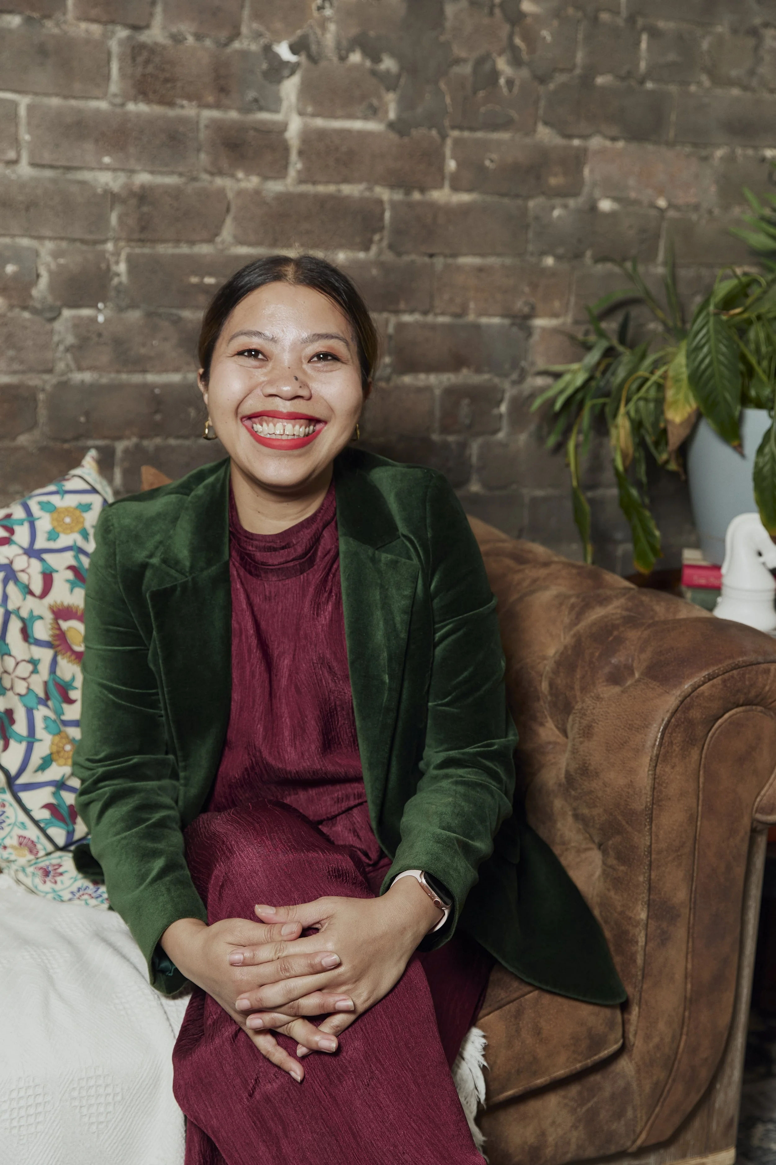 Vida Asrina Dhulst, author of The Soft Power Manual, smiling while seated on a leather sofa, wearing a green velvet blazer in front of a brick wall.