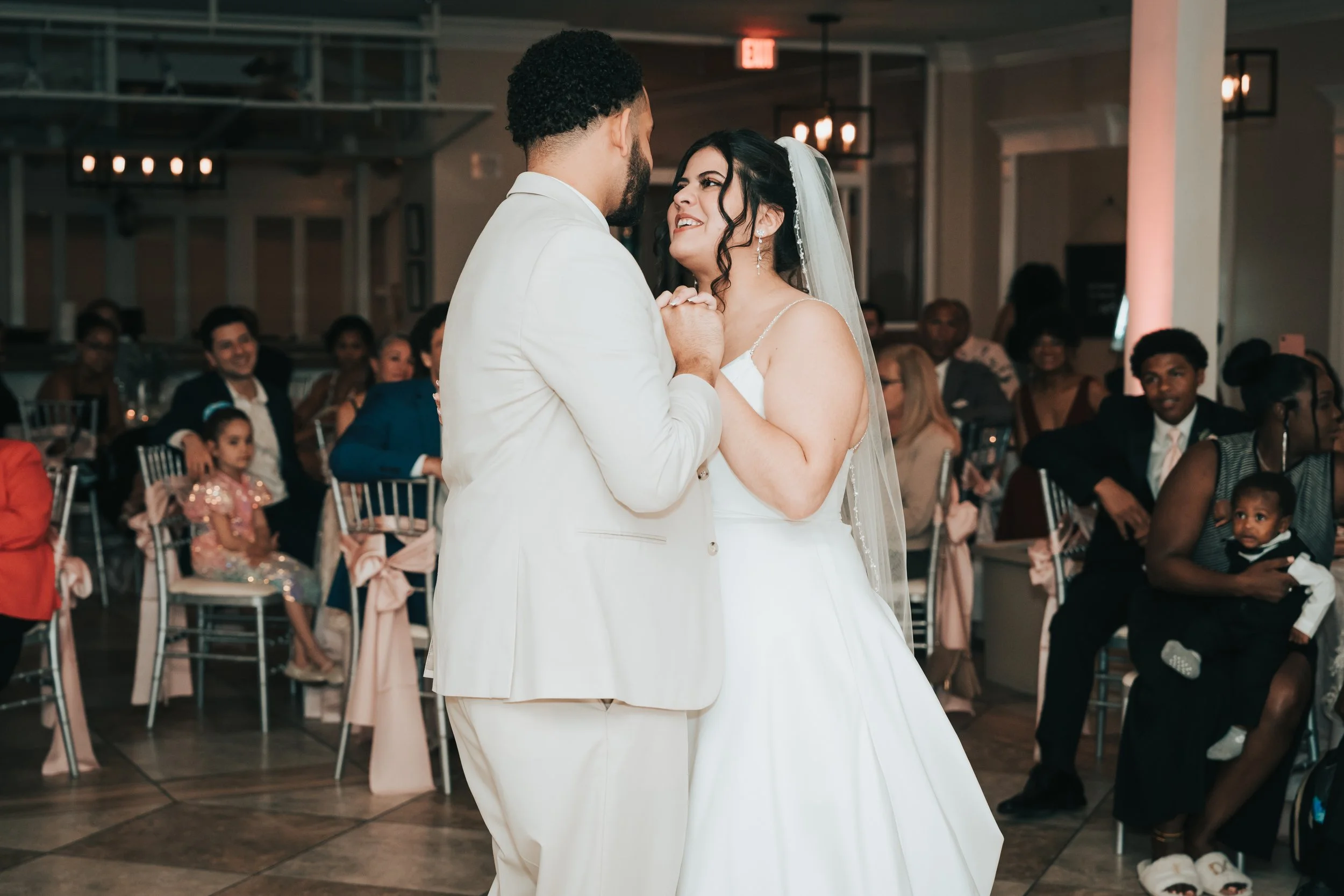 A bride and groom sharing their first dance at wedding reception with guests watching in the background.