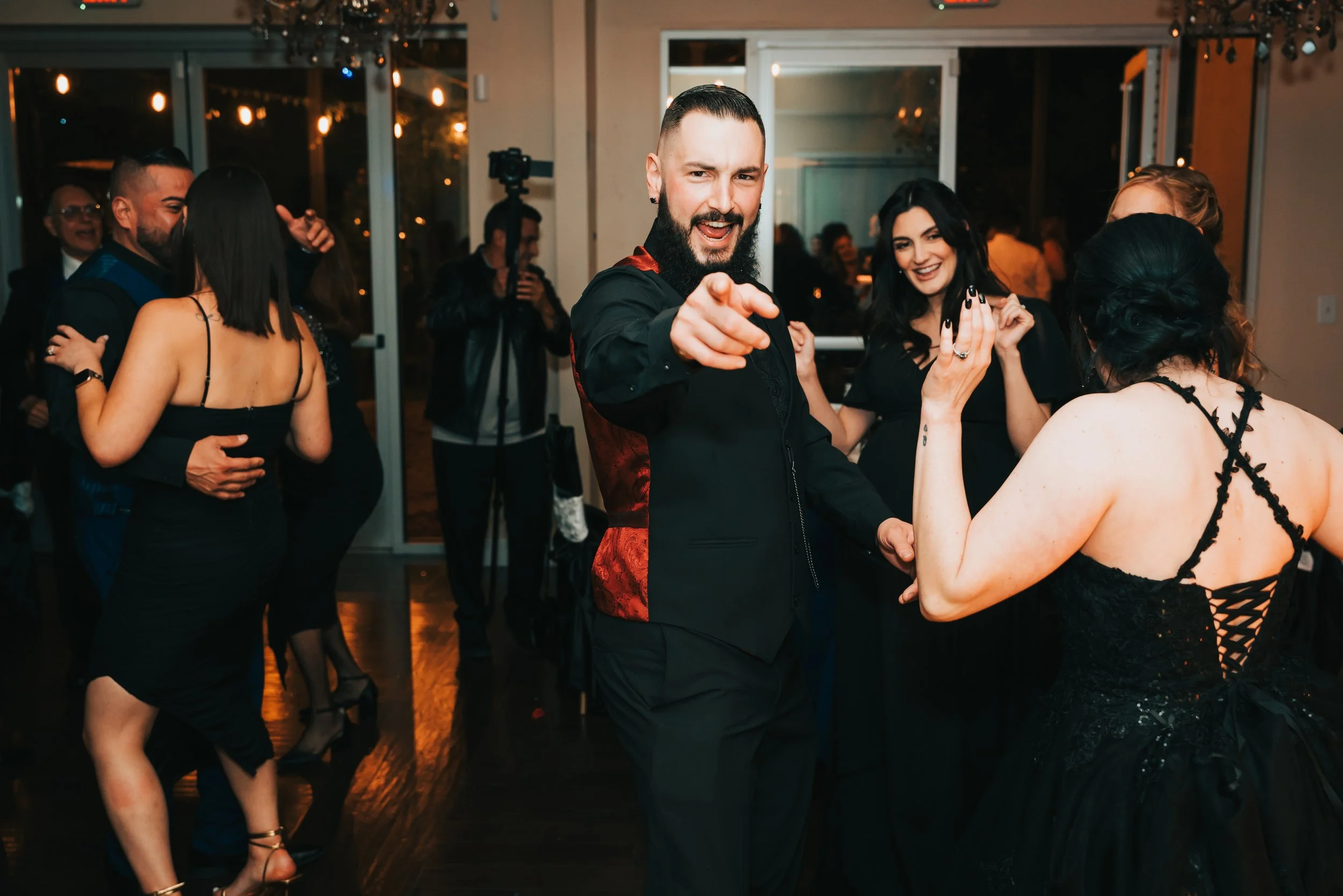 People dancing at a celebration or party, with a man in the center pointing towards the camera and smiling, wearing a black suit with a red patterned side, and women in black dresses dancing around him.