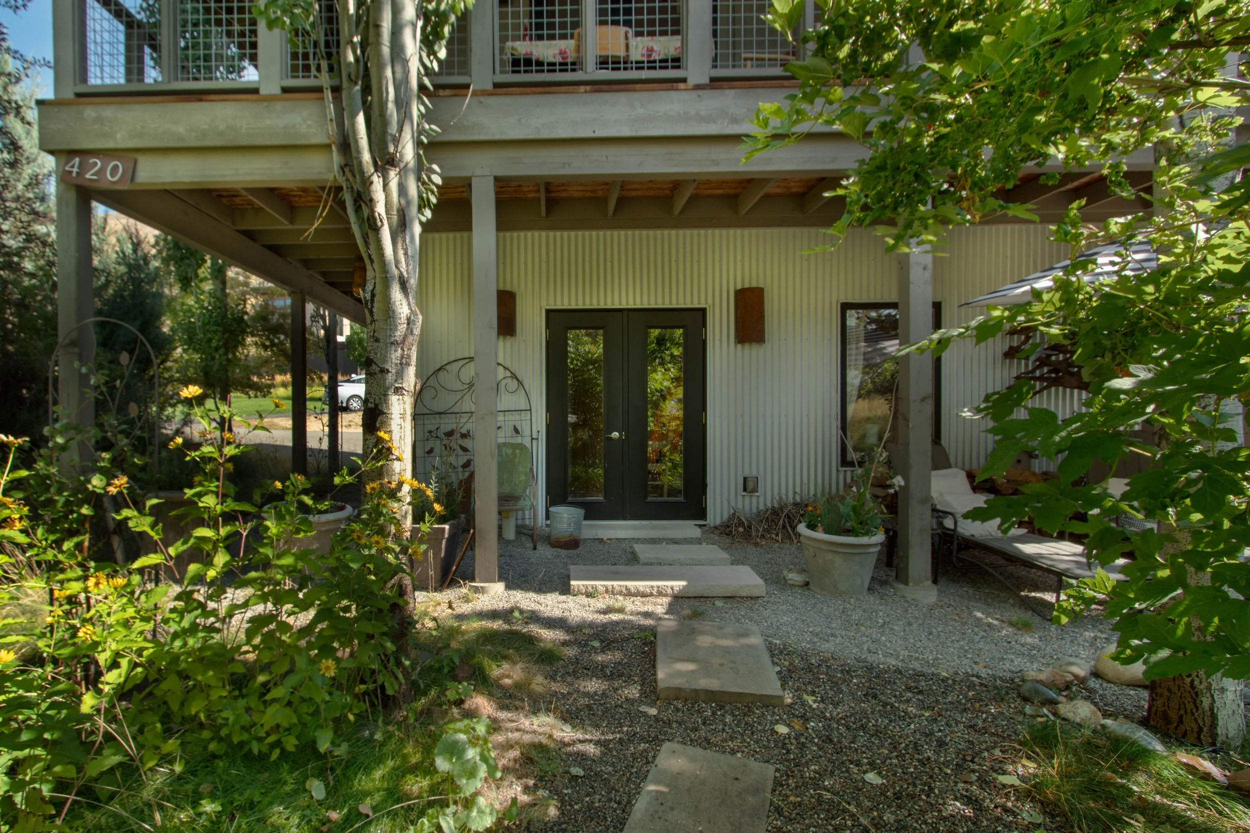 Front entrance of a modern house with a black door, concrete stepping stones, potted plants, and outdoor furniture surrounded by greenery and trees.
