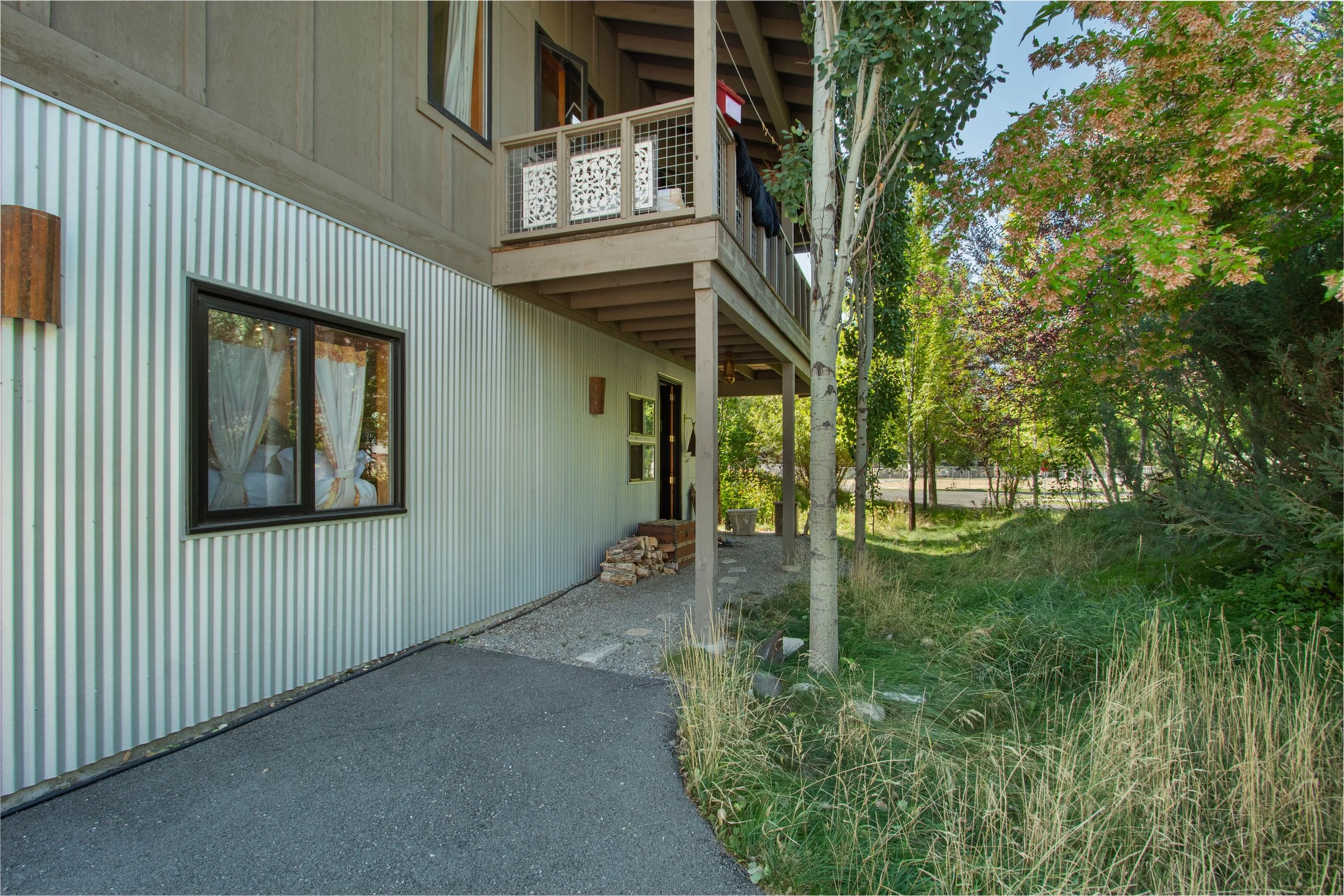 Ground level view of a modern house with a corrugated metal siding, black-framed windows, a small balcony with a railing, and a porch with a side door. There are trees and grassy areas on the right side and a paved pathway in the foreground.