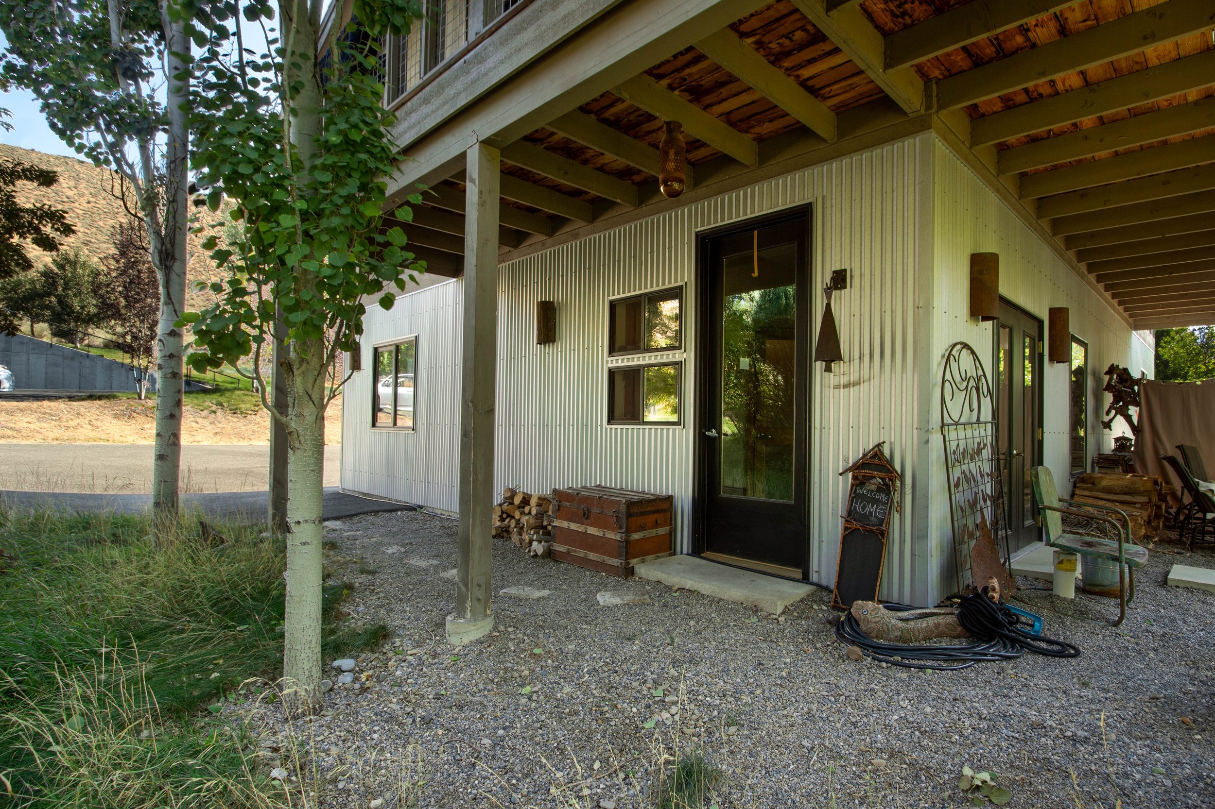 Backyard patio area of a modern home with gravel ground, outdoor furniture, woodpile, and decorative signs, under a wooden porch ceiling.
