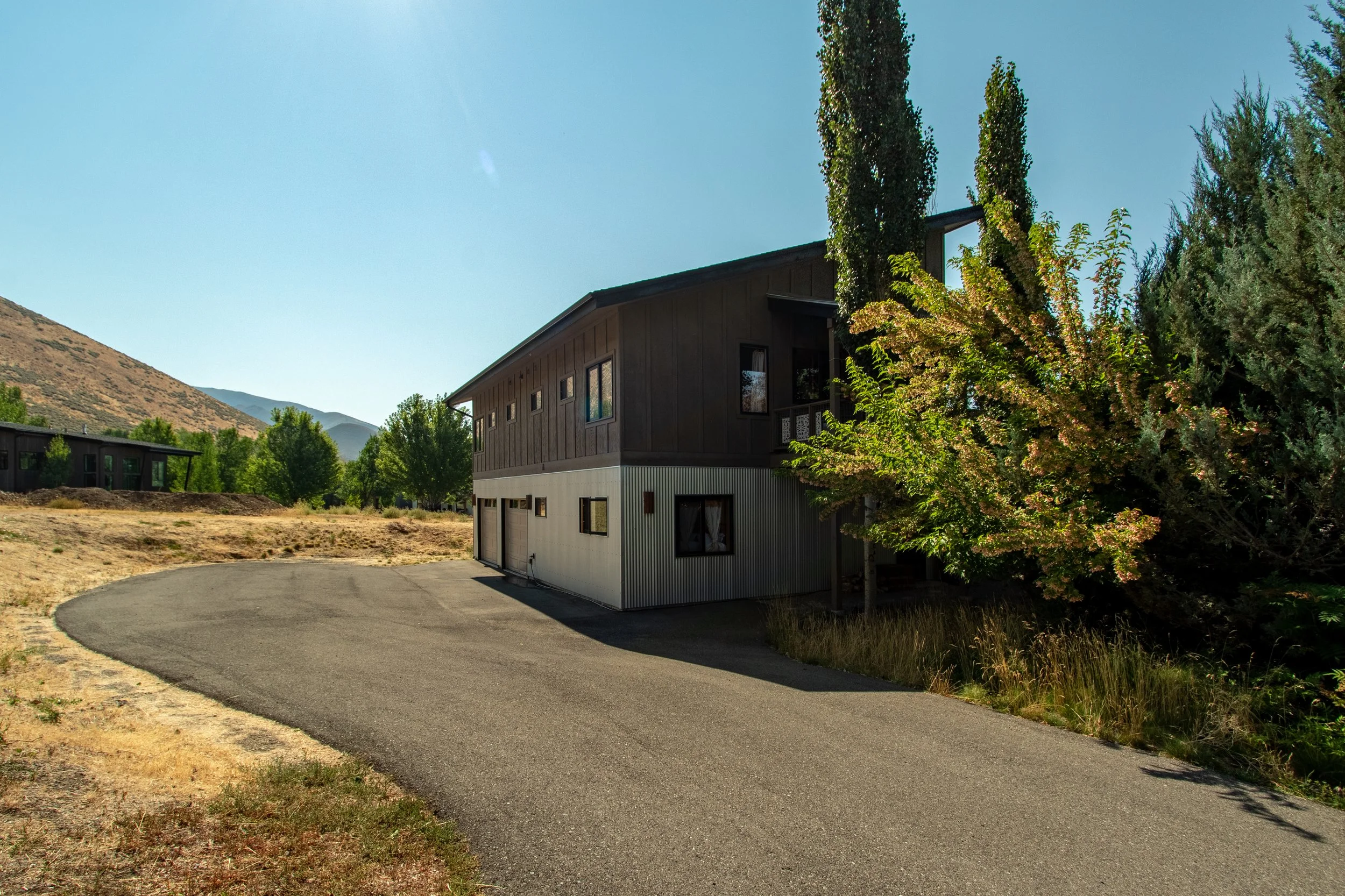 A two-story house with a dark upper level and a light lower level, surrounded by trees and dry grass, with a curved asphalt driveway leading to the house, set in a rural area with hills in the background.