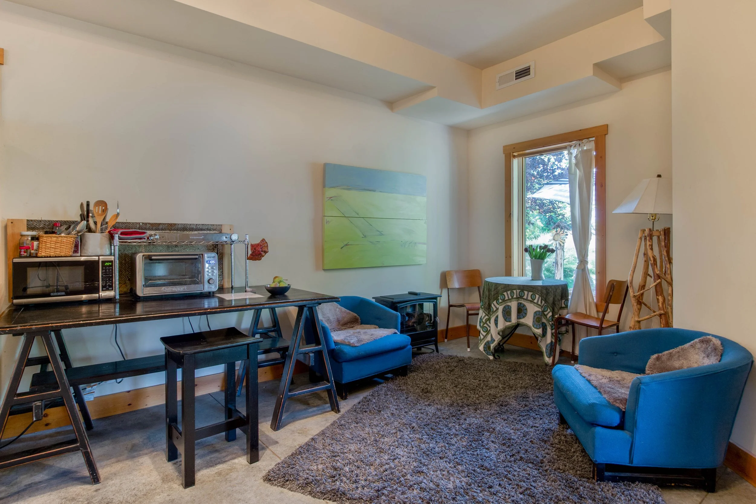 A cozy living room with two blue chairs, a small round dining table with a green patterned tablecloth, a large window with curtains, a lamp, a painting on the wall, a dark brown rug, a black side table, and a black countertop with kitchen appliances.