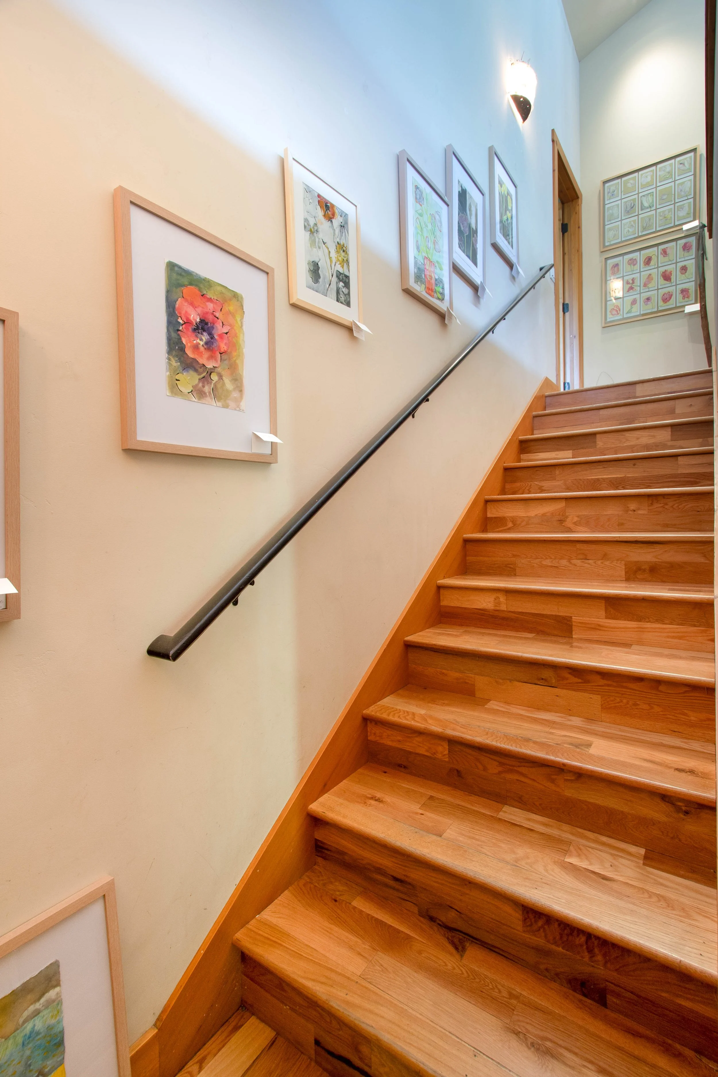 A wooden staircase with a black handrail along a cream-colored wall decorated with framed artwork, leading to an upper floor with additional framed art on the walls.