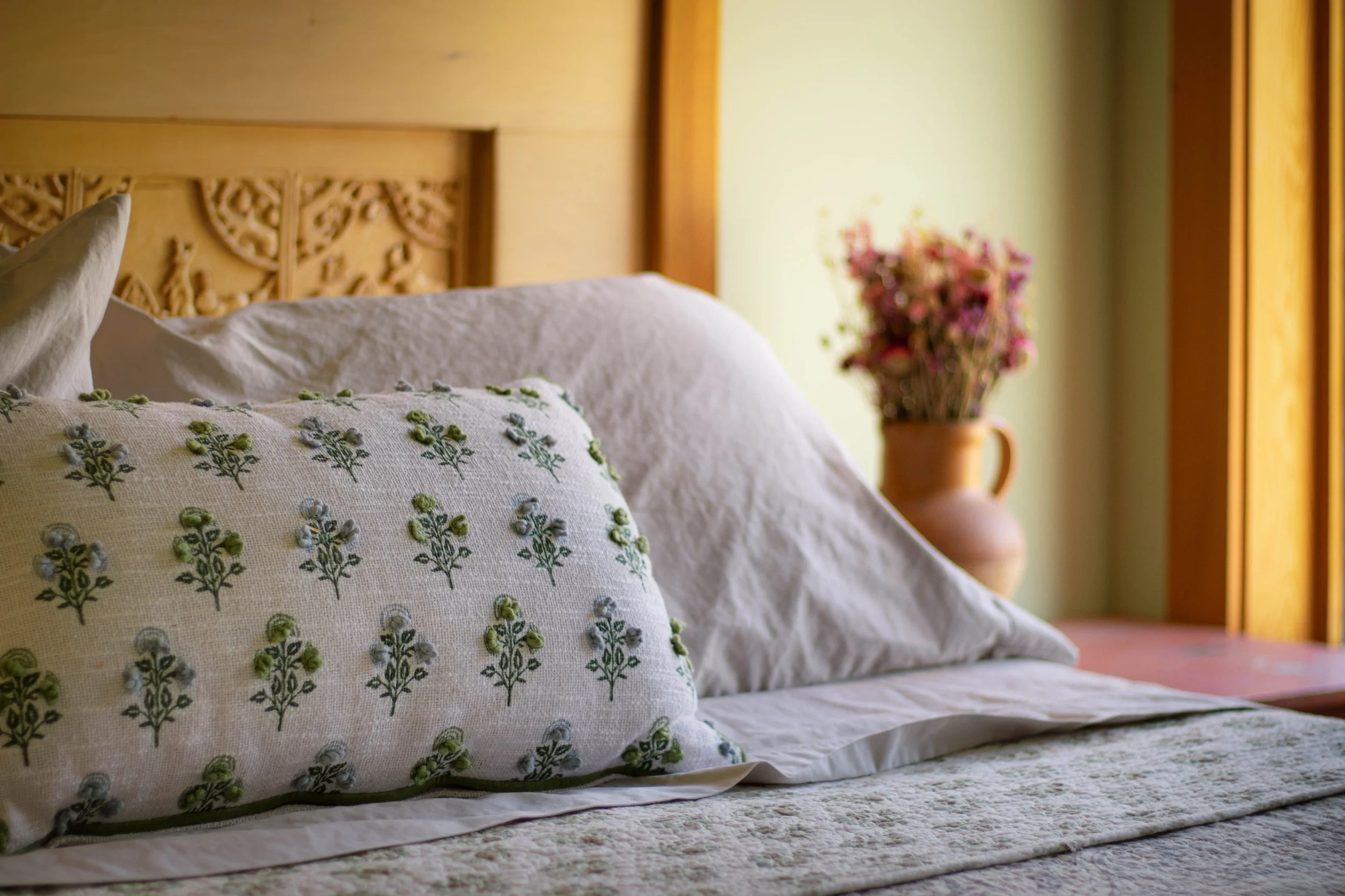 A neatly made bed with embroidered pillows and a textured bedspread, in a room with a wooden headboard and a vase of dried flowers on the side table.