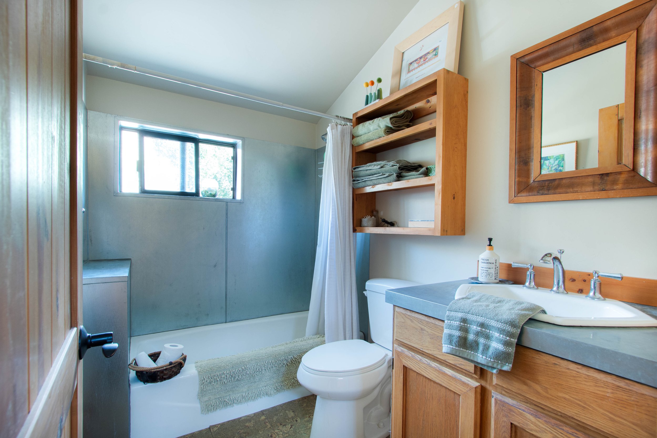 A small bathroom with a shower, toilet, and vanity with a mirror. The shower has a window, and there are shelves with folded towels and decor. The sink has soap and a hand towel.