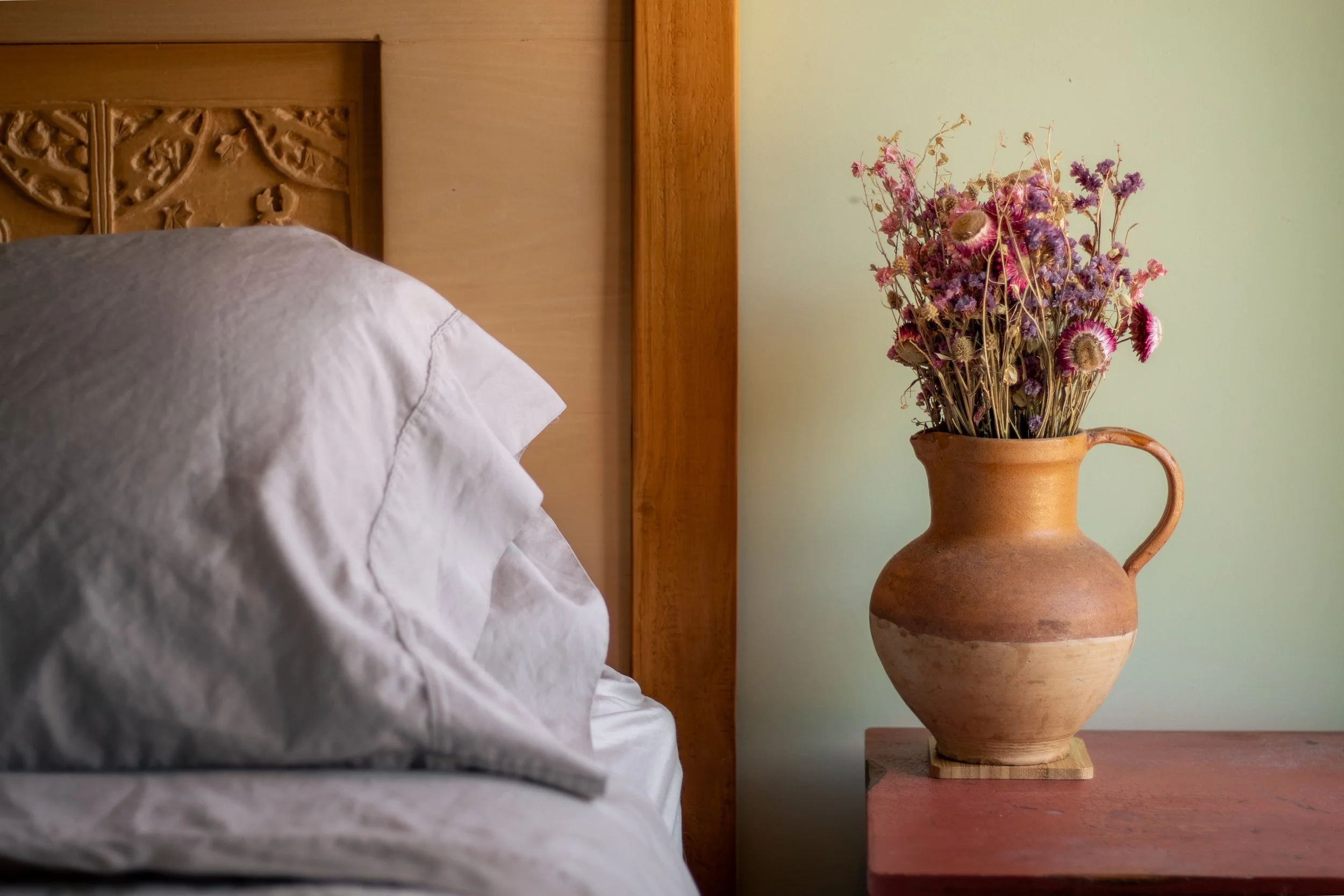 A rustic brown terracotta vase with a handle filled with dried purple and pink flowers, placed on a reddish wooden table beside a bed with a white pillow, against a pale green wall.