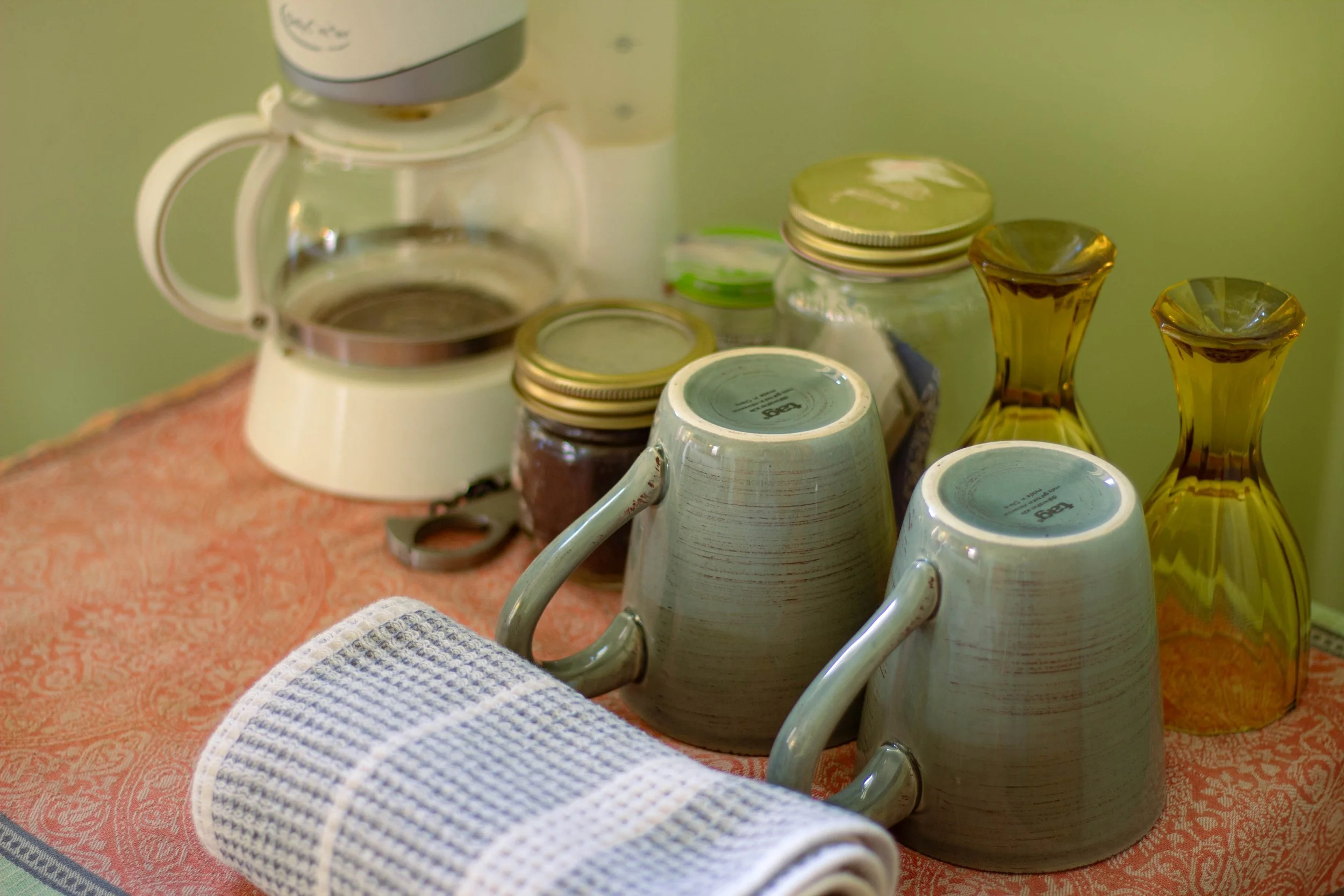 Coffee cups, glass vases, jars, a coffee pot, and a rolled towel on a table with an orange patterned tablecloth.