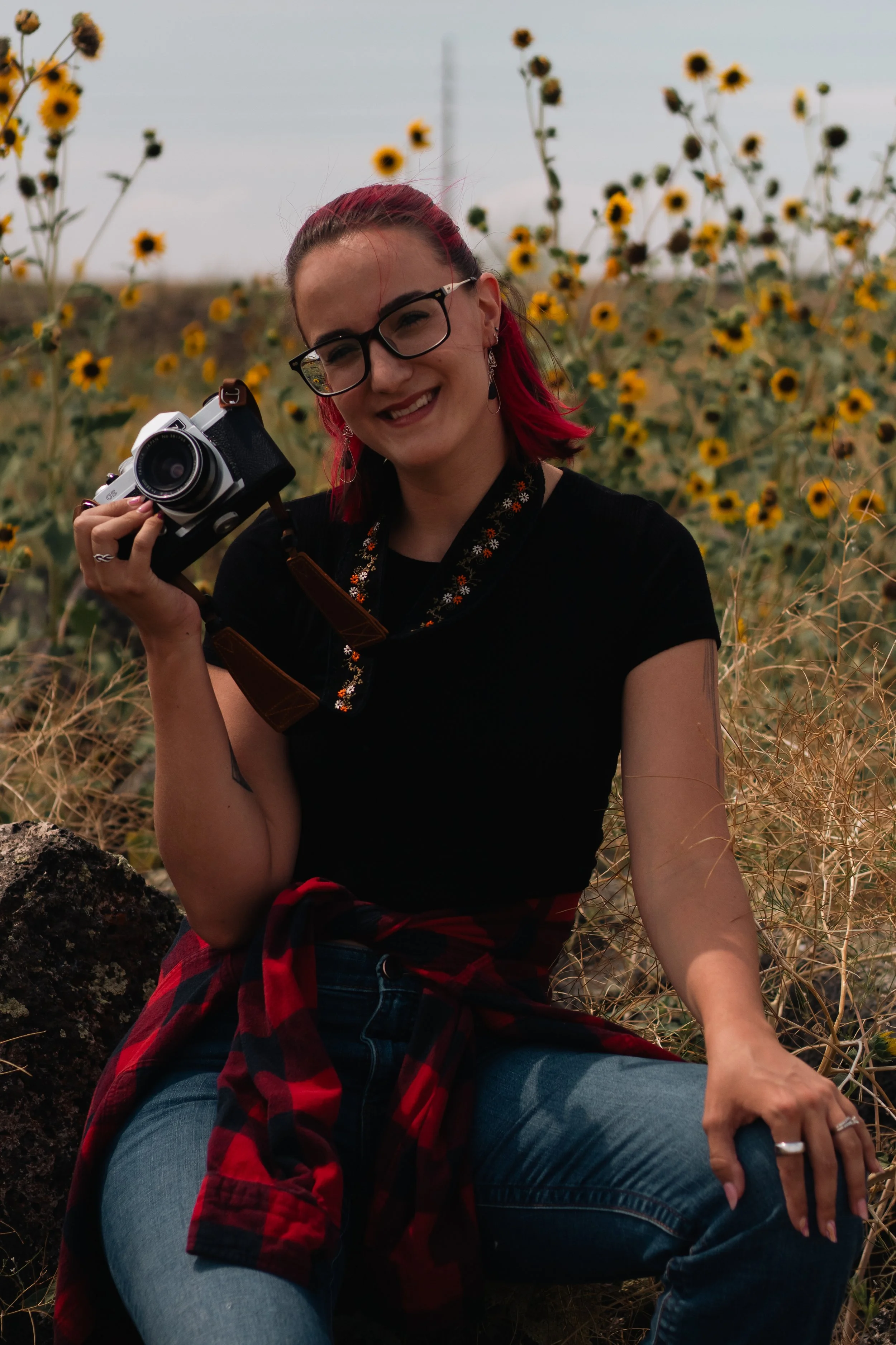 A young woman with red hair, glasses, and earrings sitting outdoors among sunflowers, holding a camera, smiling, wearing a black shirt and a red and black flannel tied around her waist.
