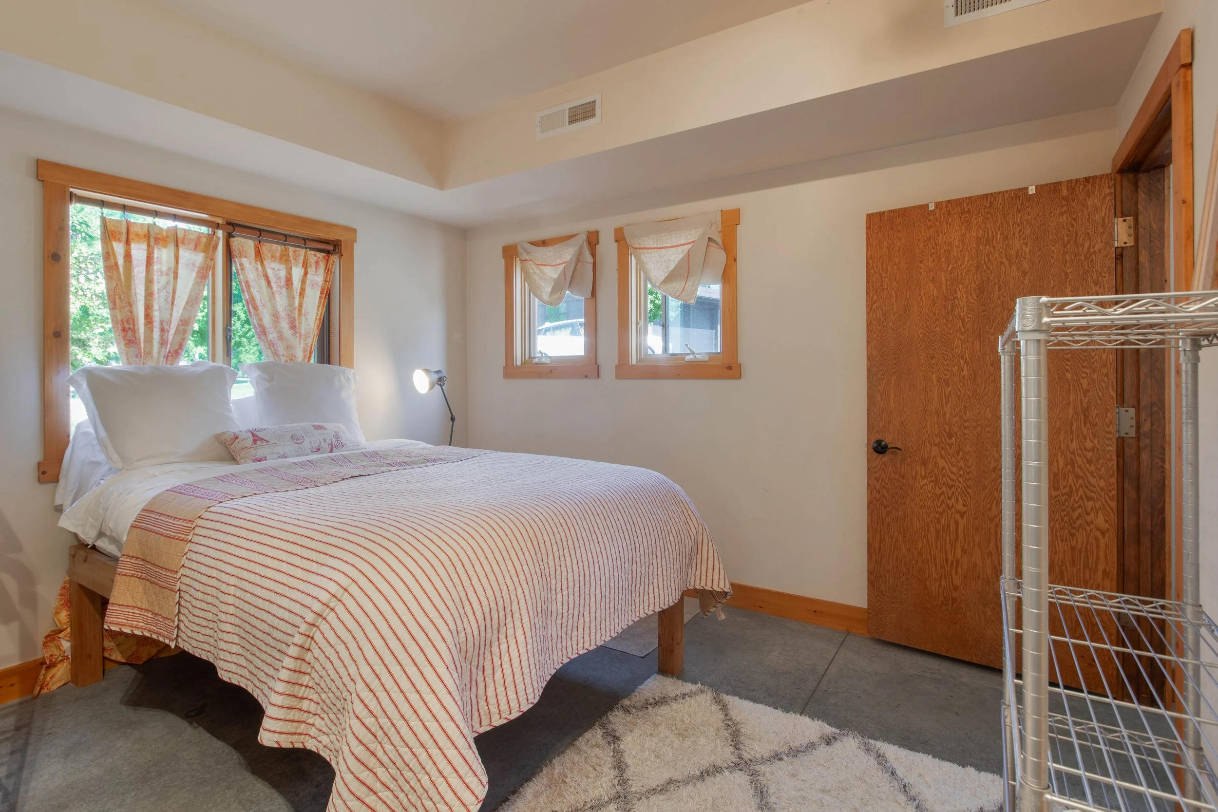 Bedroom with a wooden bed frame, white bedding with red stripes, two large pillows, a smaller patterned pillow, wall-mounted reading lamp, three windows with curtains, wooden trim, beige walls, a door, and a white shelving unit.