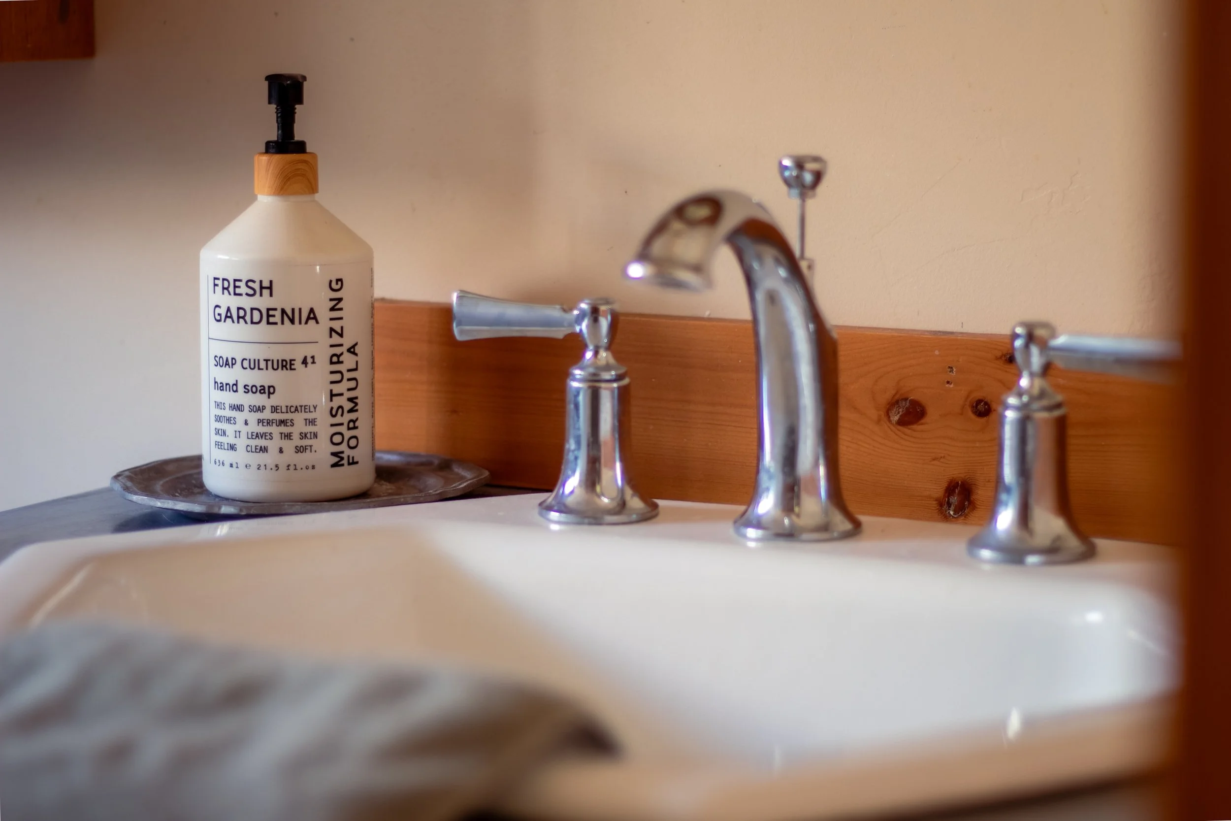 A bathroom sink with a chrome faucet and handles, a wooden backsplash, and a bottle of hand soap on a silver tray.