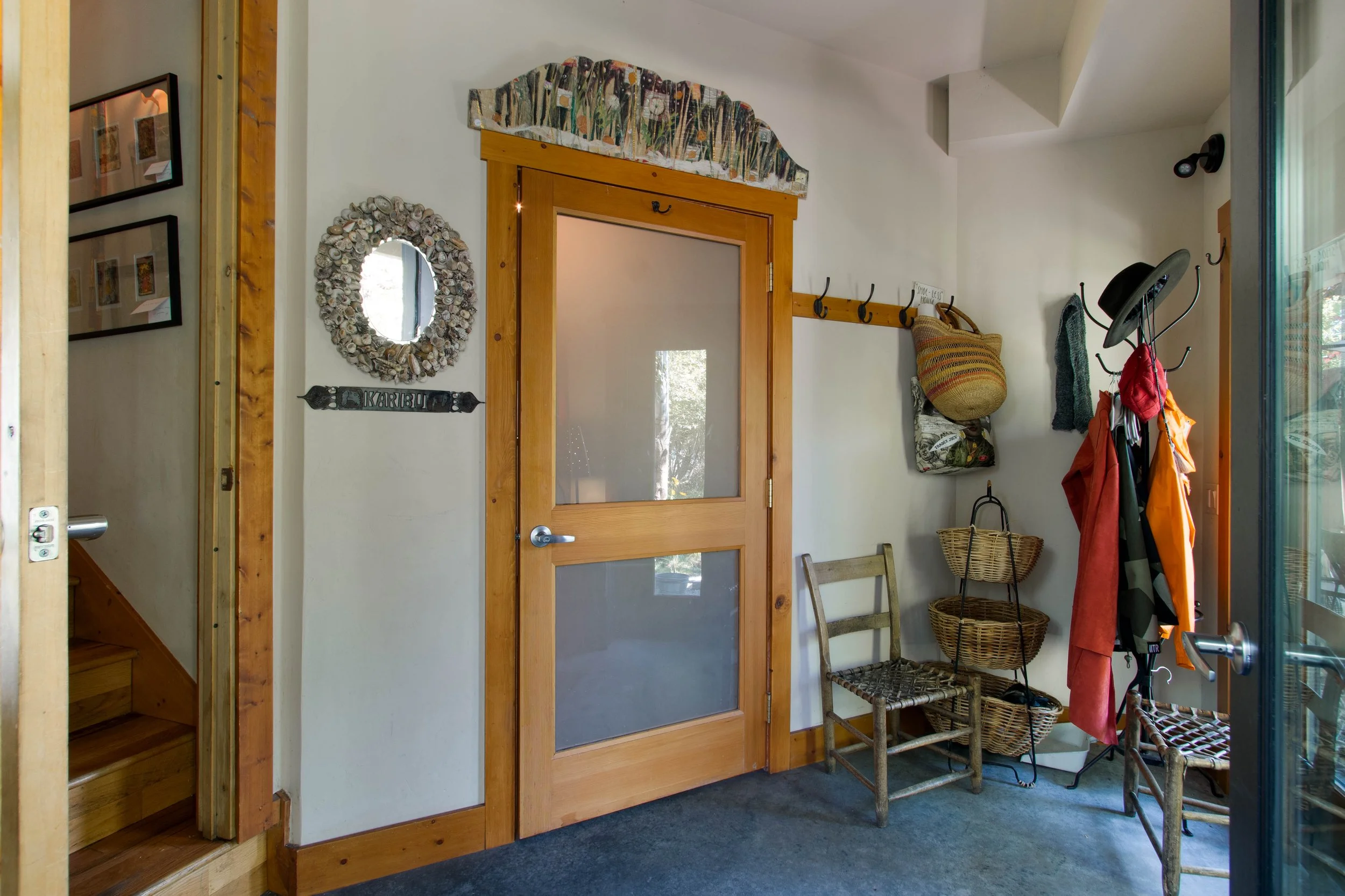 Interior of a house entryway with a wooden door featuring frosted glass, a wall-mounted coat rack holding various jackets, hats, and bags, a small wooden chair, baskets, a mirror decorated with seashells, and framed pictures on the wall.