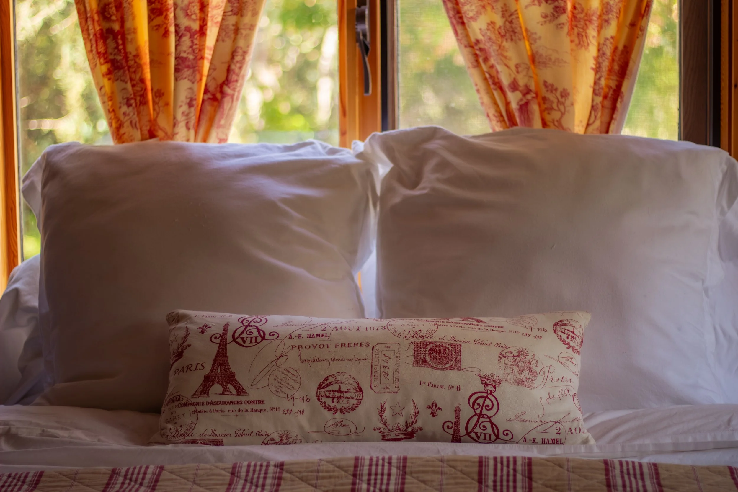 A bed with two large white pillows and a decorative pillow with a Paris-themed design, in front of a window with floral curtains and greenery outside.