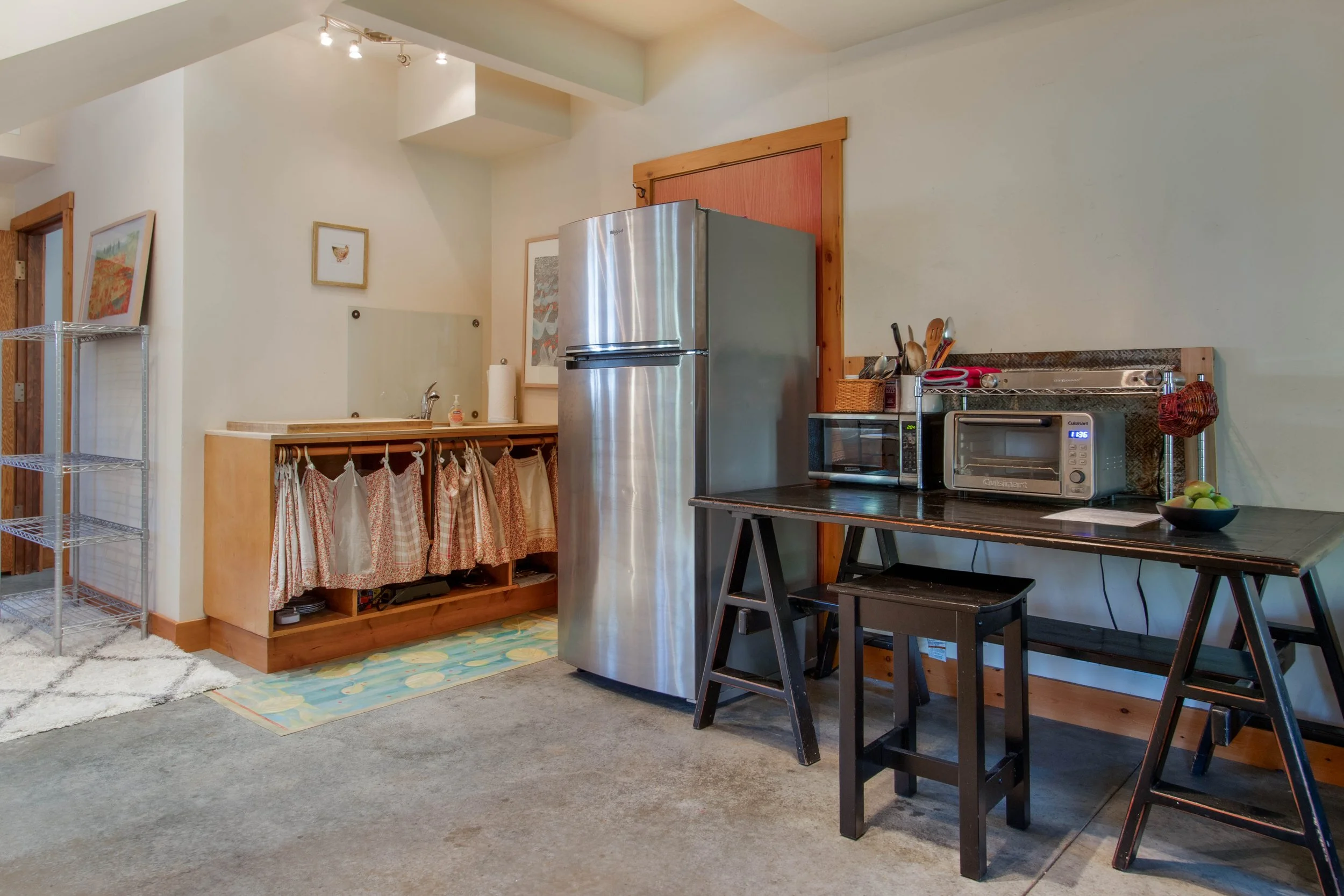 A kitchen with a stainless steel refrigerator. To the right, there is a black table holding small appliances including a microwave oven, a toaster oven, and a microwave. There are wooden stools near the table. In the background, a sink with a paper t