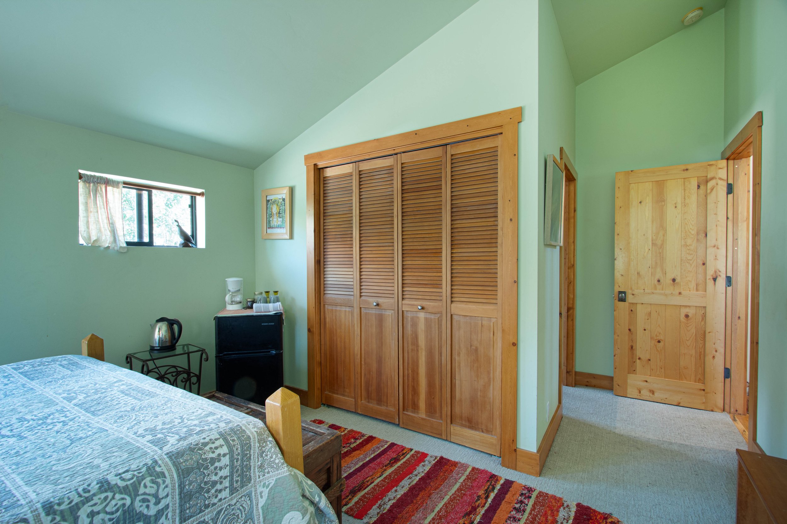 Comfortable bedroom with green walls, wooden closet doors, a window with a curtain, a bed with a patterned bedspread, a small black refrigerator, a coffee maker, a kettle, a small table, and a colorful striped rug.