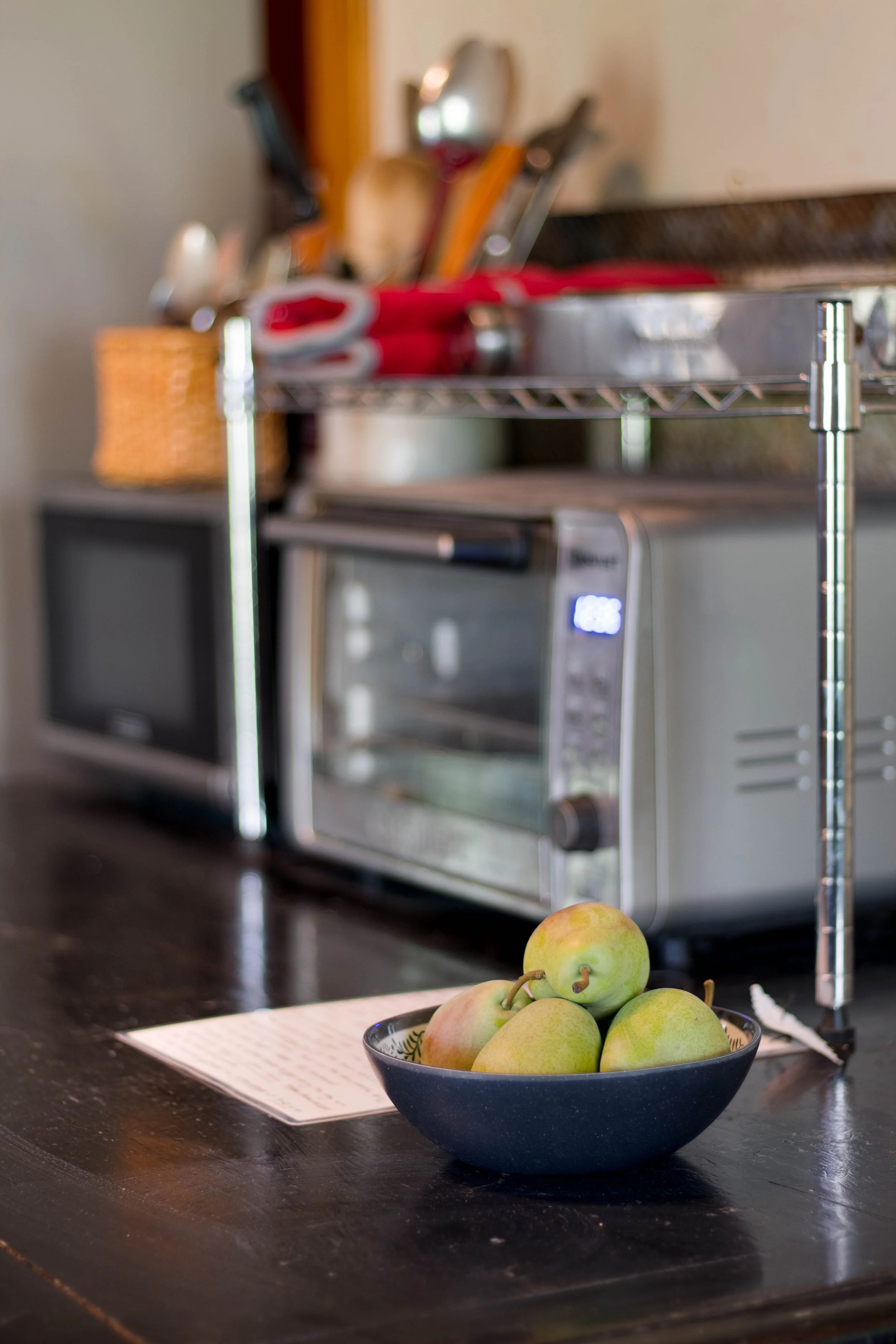 A bowl of pears on a dark wooden kitchen countertop in front of a microwave, with a metal rack holding utensils and a toaster in the background.