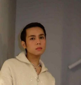 A woman with short dark hair wearing a cream-colored hoodie and a necklace, standing indoors against a plain wall.