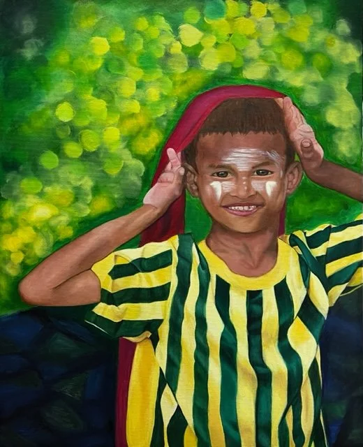 A young boy with a big smile wearing a yellow and green striped shirt, holding a dark red cloth over his head, standing outdoors against a green leafy background.