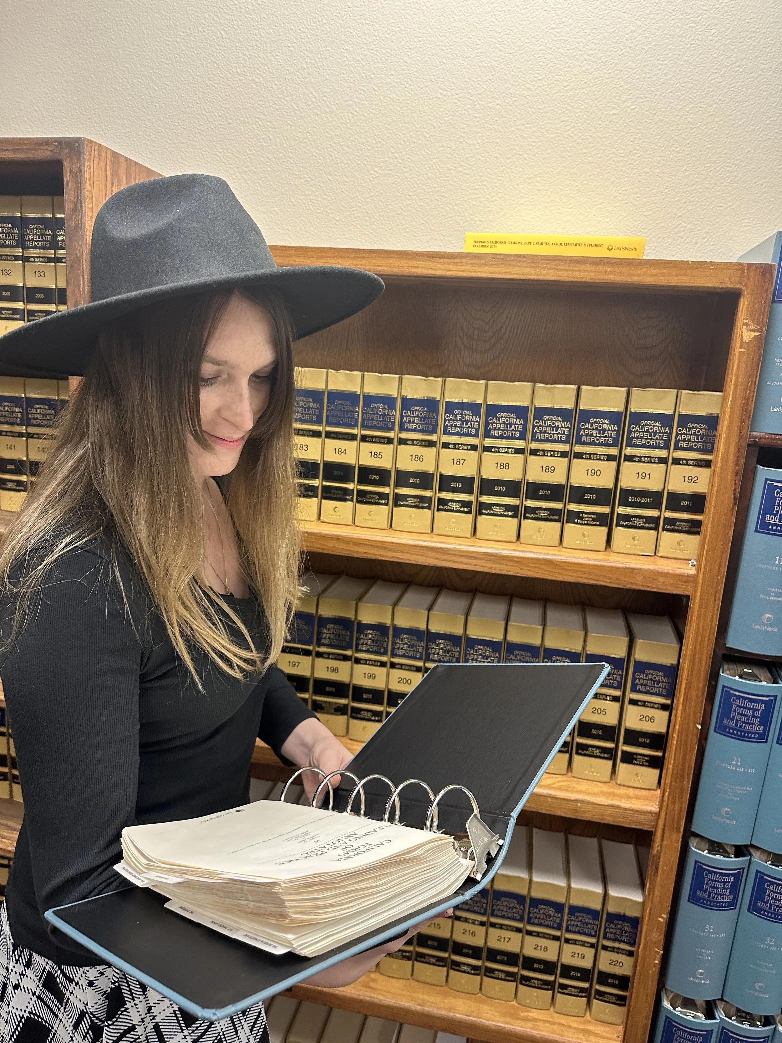 A woman with long brown hair, wearing a large gray hat and black top, looks at a black folder with legal documents on it, in front of shelves filled with legal case reports.
