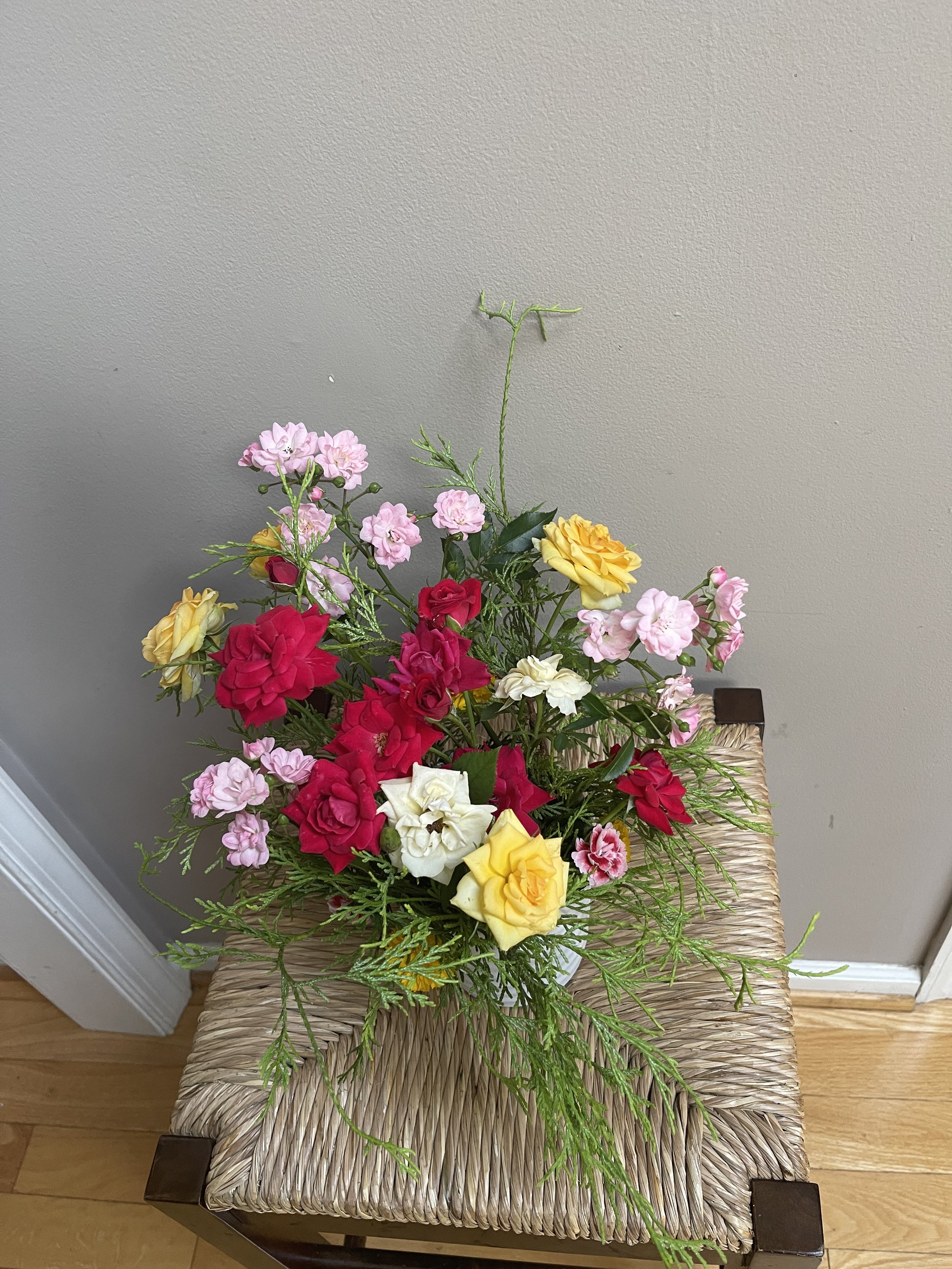A floral arrangement with pink, yellow, red, and white flowers on a woven seat stool against a neutral wall and hardwood floor.