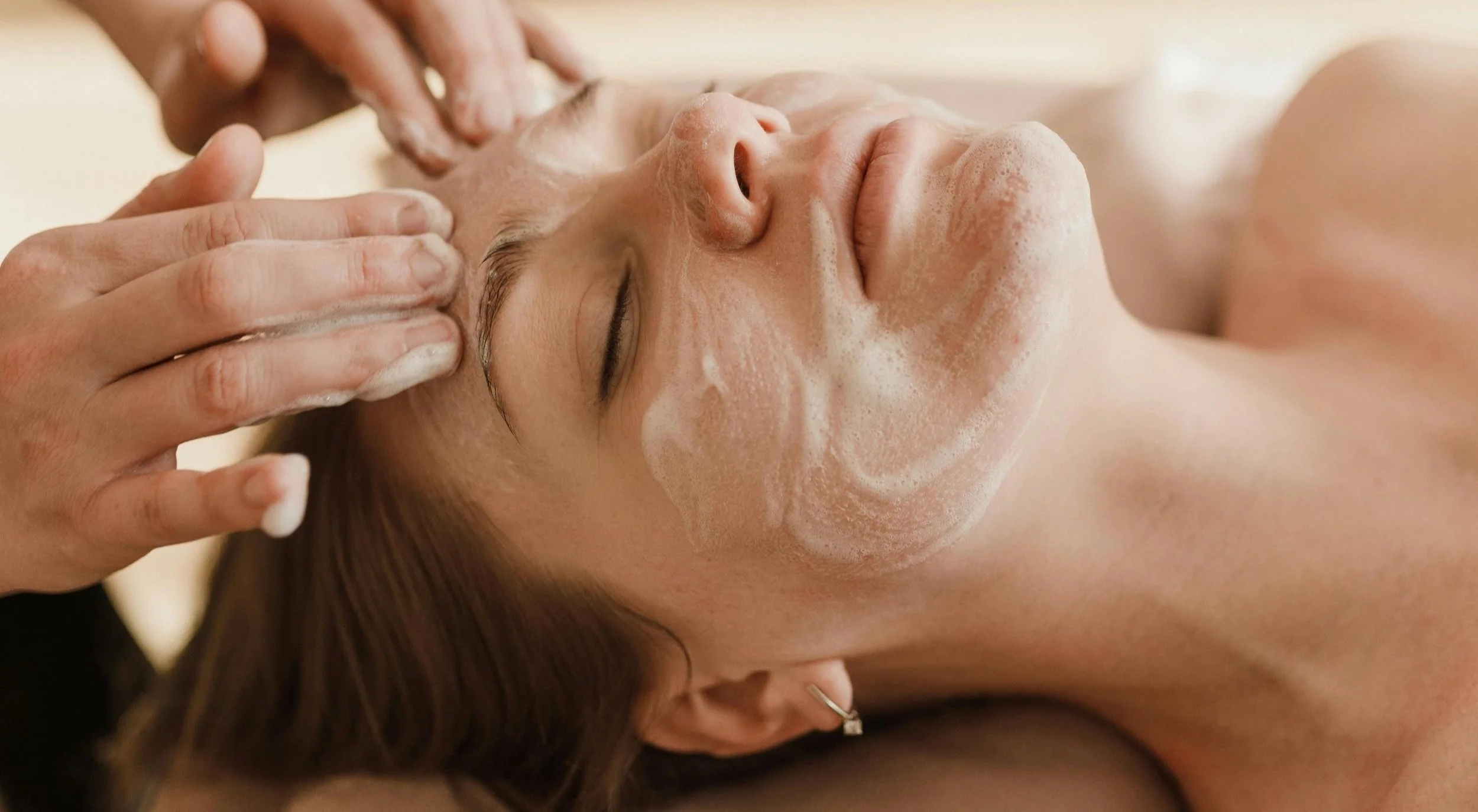 A woman lying down receiving a facial massage with soap or foam on her face.