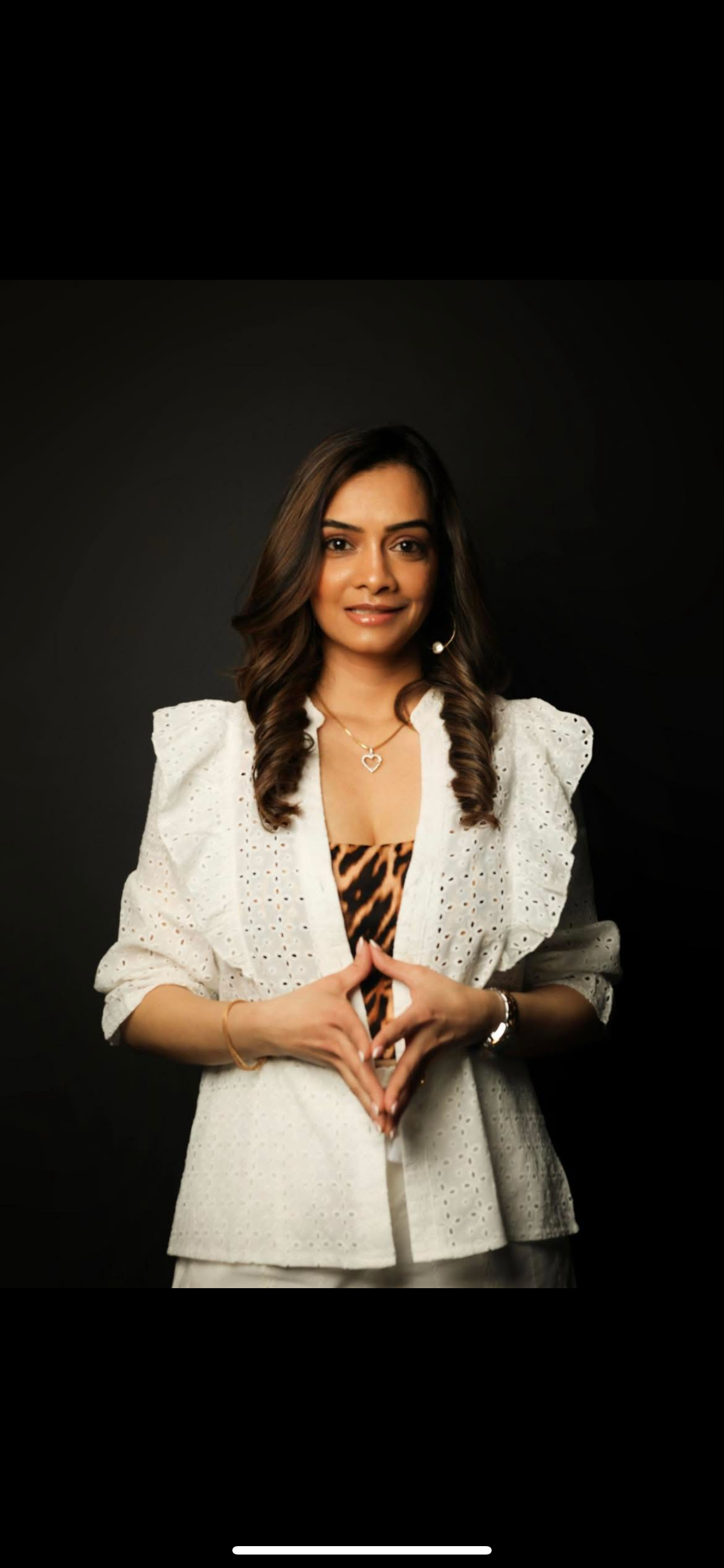 A woman with shoulder-length wavy brown hair, wearing a white eyelet blazer over a leopard print top, posing against a black background, making a heart shape with her hands.