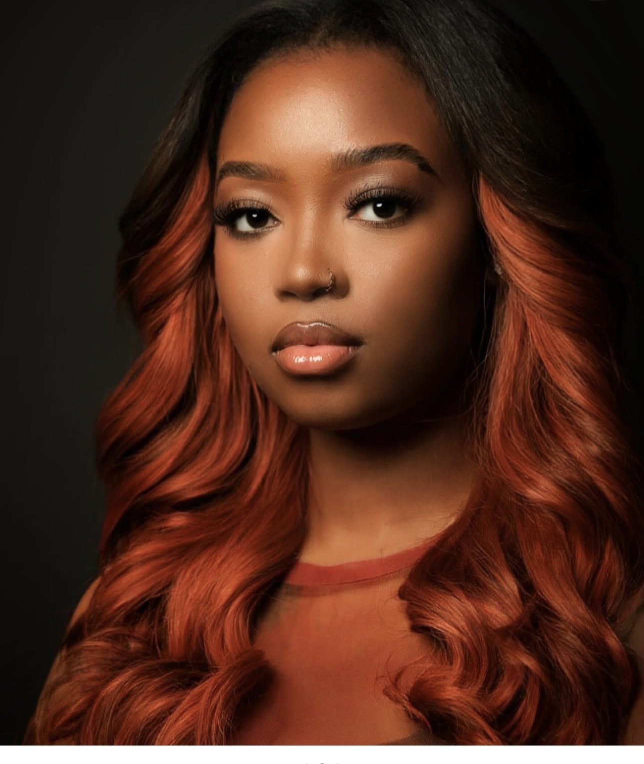 A portrait of a young African American woman with long wavy hair that transitions from black at the roots to bright orange at the ends, against a dark background.