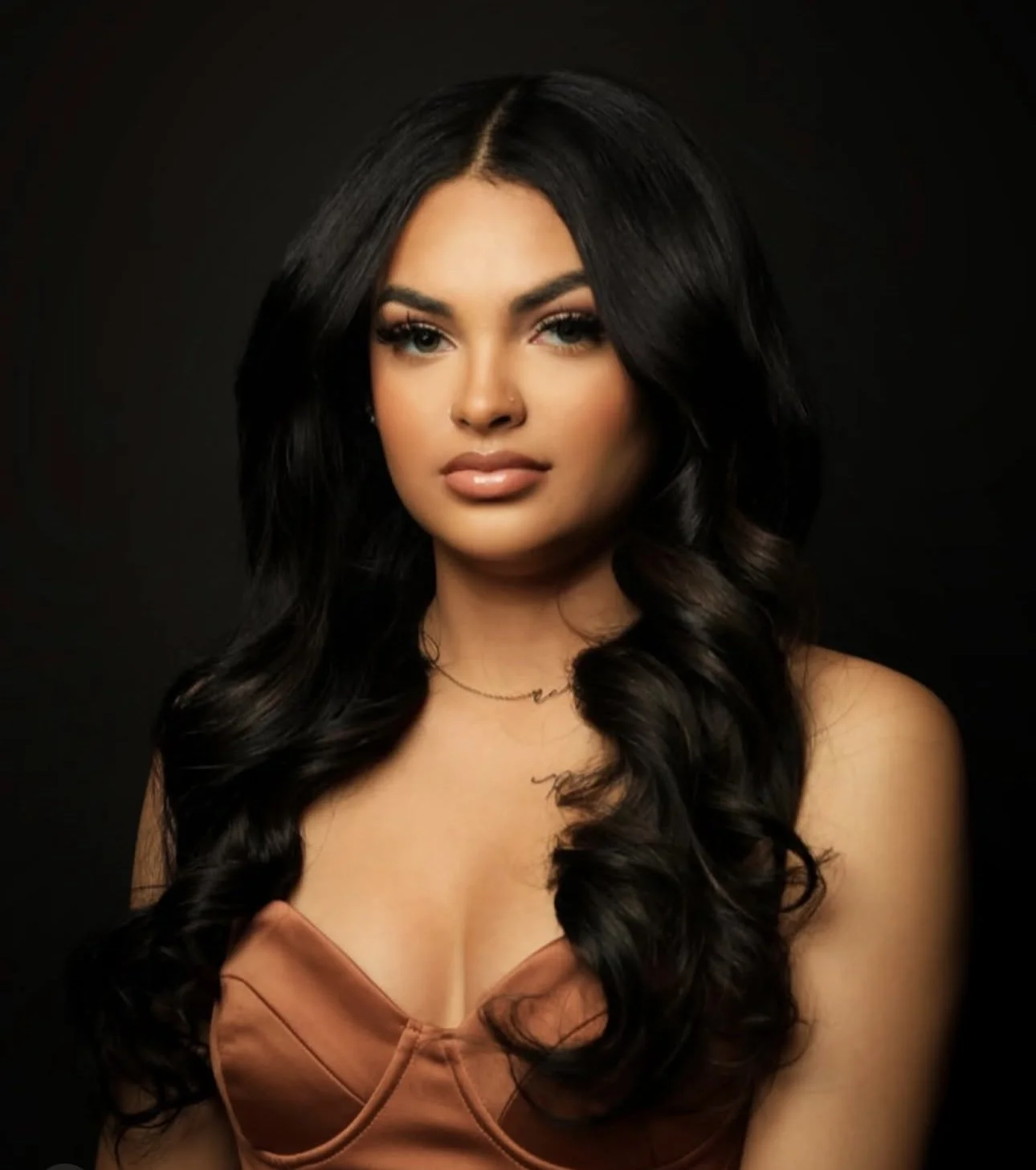 A young woman with long black wavy hair, wearing a tan top and a delicate necklace, posing against a dark background.