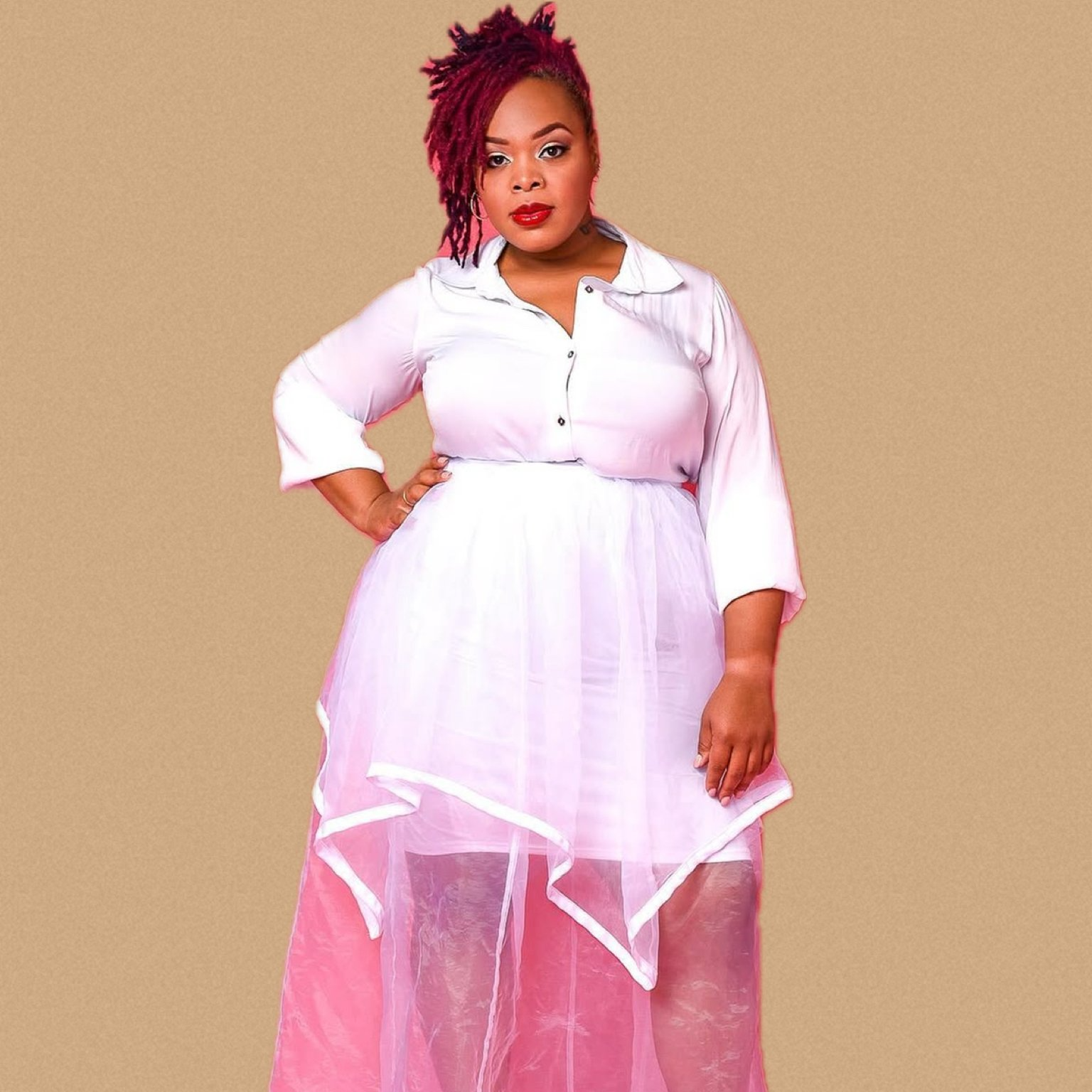 A woman with short, reddish-dyed dreadlocks wearing a white button-up dress with a sheer pink overlay, standing against a plain brown background.