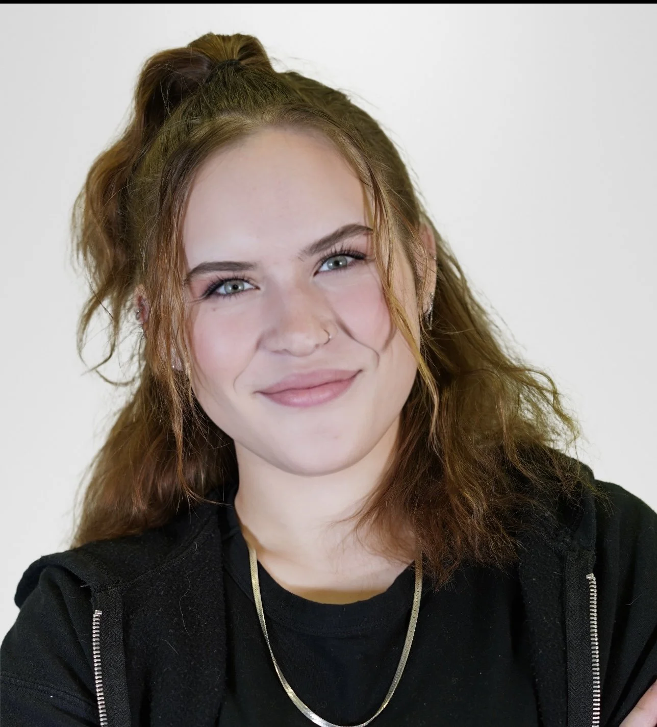Young woman with light skin, wavy red hair tied in a half ponytail, light blue eyes, and a small nose ring, smiling against a plain white background.