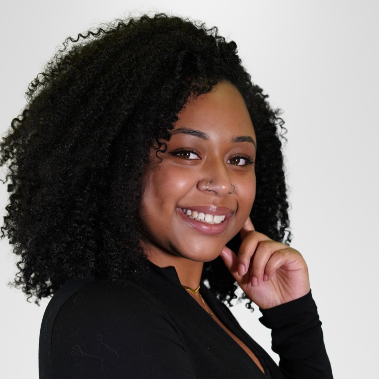 A young woman with curly black hair, smiling with her teeth showing, wearing a black top and a small gold necklace, with a neutral white background.