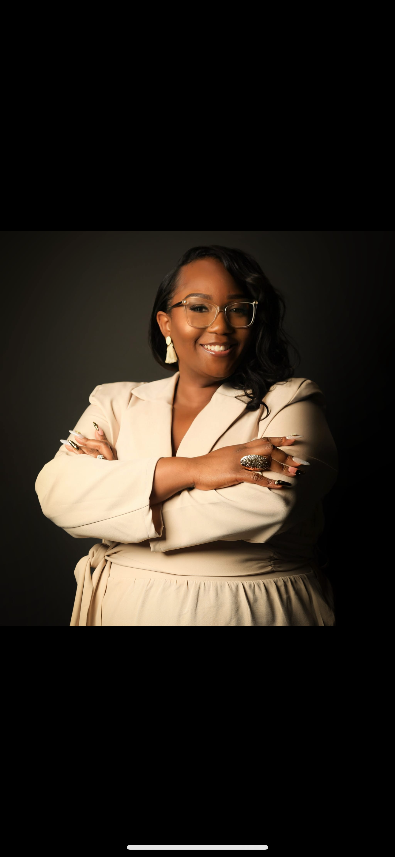 A confident woman with glasses and earrings smiling at the camera, crossing her arms, dressed in a cream-colored blazer and skirt, standing against a dark background.