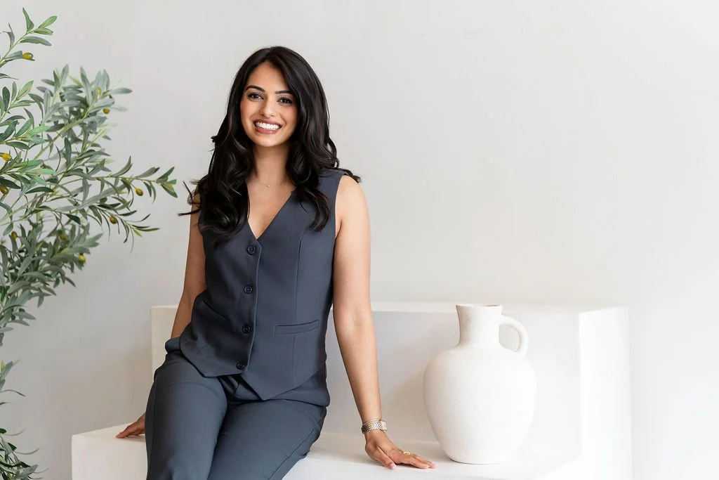 A woman in a dark gray sleeveless vest and matching pants, smiling while sitting on a white surface next to a white ceramic vase, with a plant in the background.