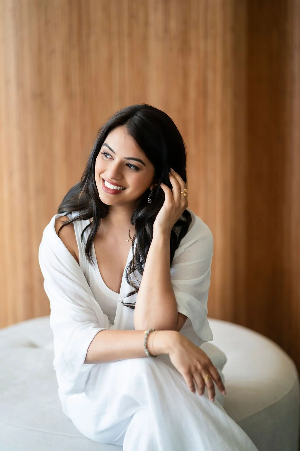 Dr. Komal Yadav with black hair, wearing a white dress, smiling and touching her hair, sitting in front of a wooden wall.