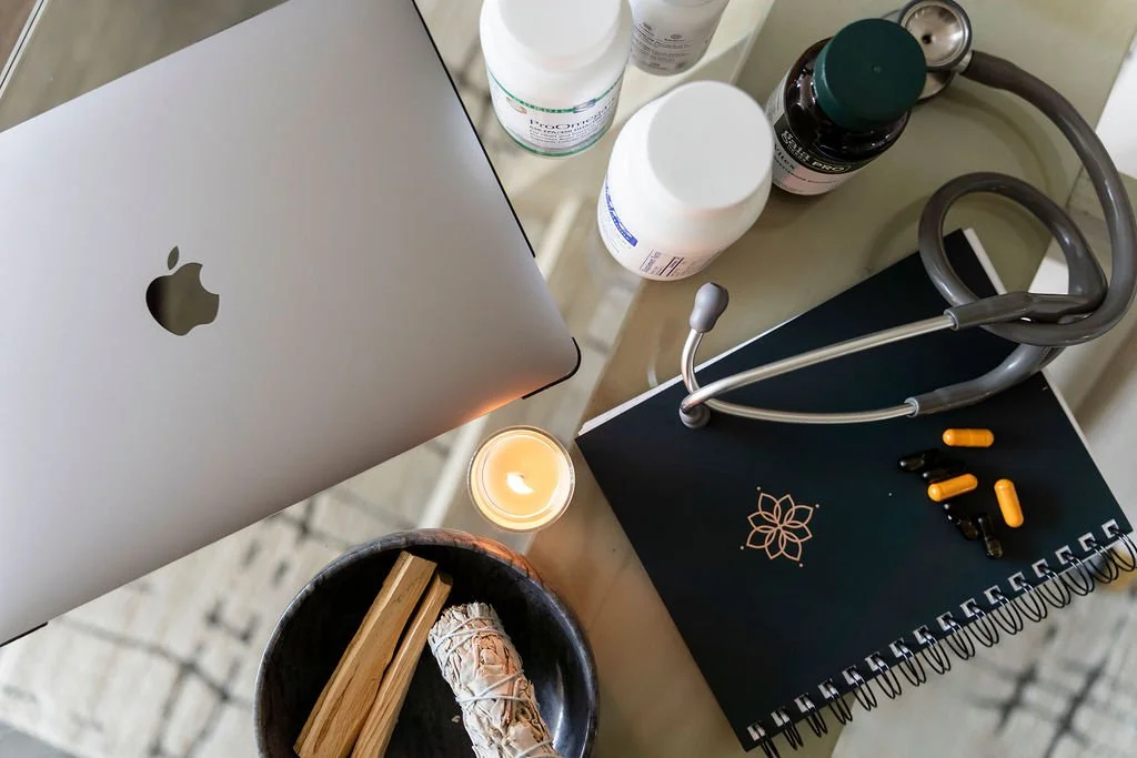 A workspace with a MacBook, bottles of supplements, a stethoscope, pills, a candle, and a black notebook with a flower logo.