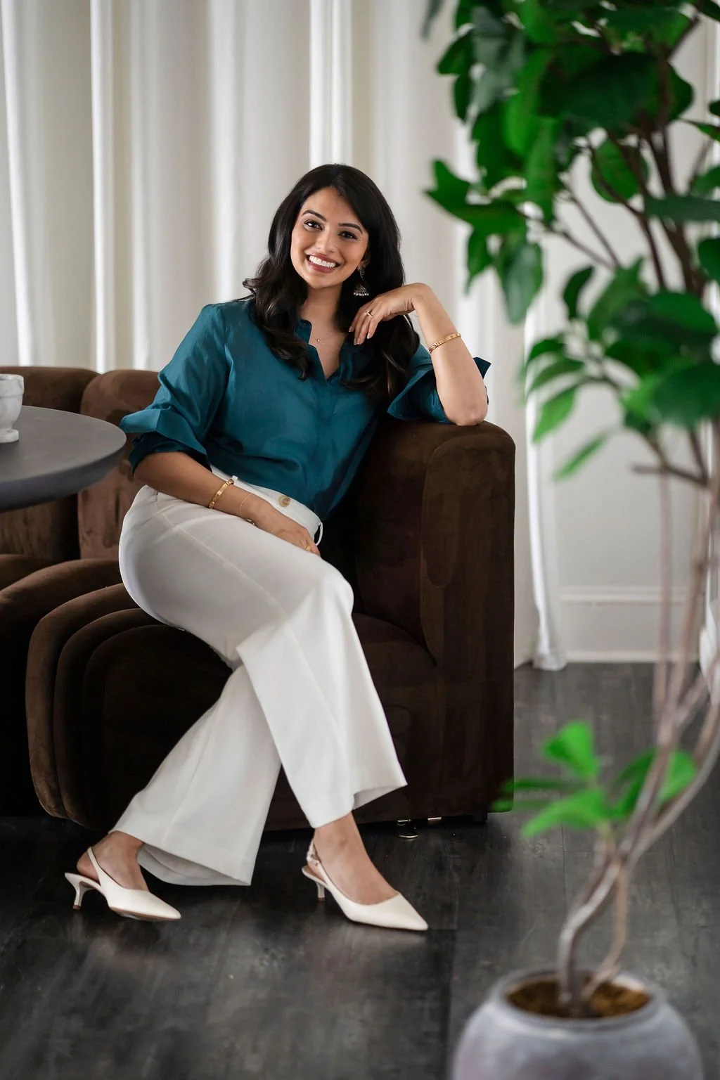 A woman with long dark hair, smiling, sitting on a brown sofa in a well-lit room with white curtains, green plants, and a dark wood floor. She is wearing a teal blouse, white wide-leg pants, white heels, and jewelry.