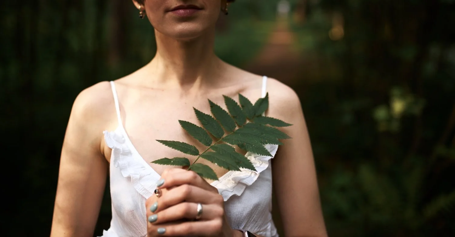 A woman in a white dress holding a large green leaf in front of her chest in a forest setting.