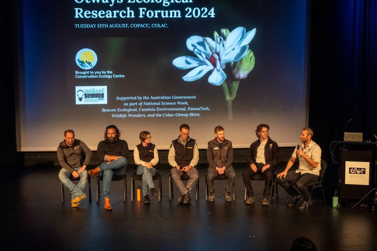 a panel of 7 speakers on a stage sitting down with a presentation on a screen behind them