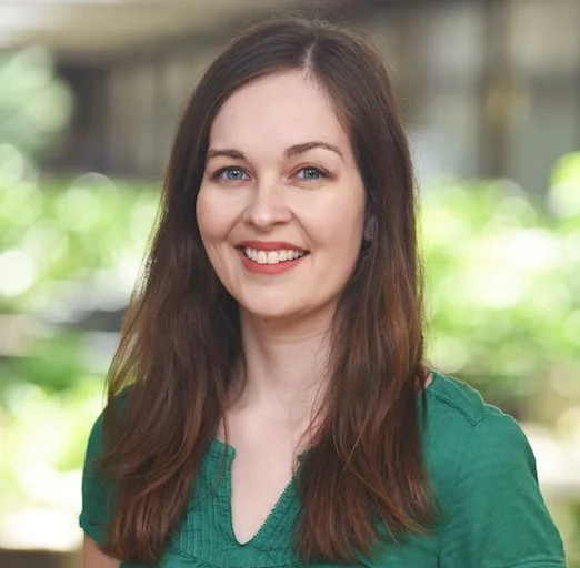 A woman with long brown hair and blue eyes smiling outdoors with green blurred background.