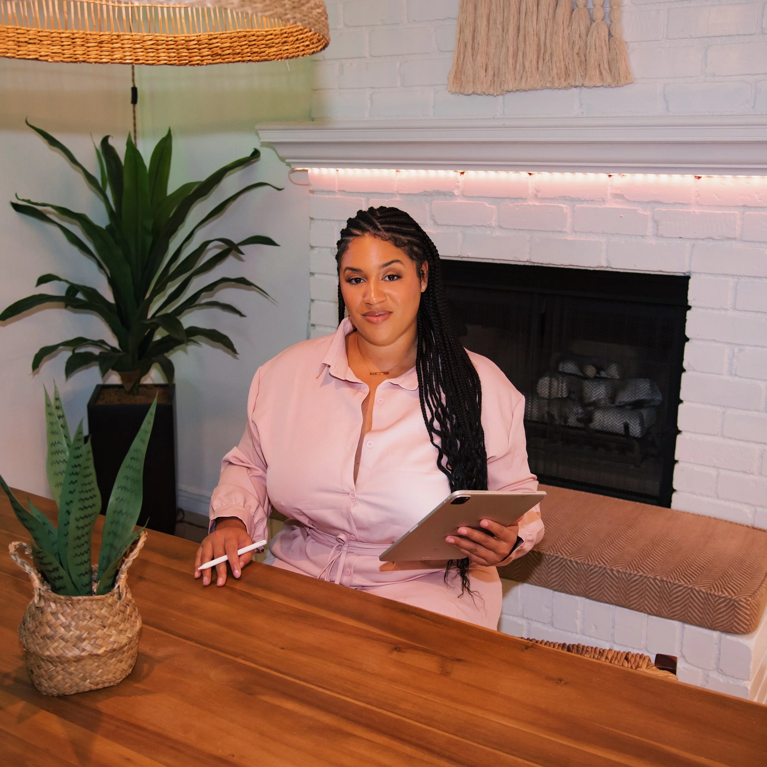 A woman sitting at a wooden table holding a tablet and stylus in a cozy room with a white brick fireplace, green plants, and decorative wall hanging.