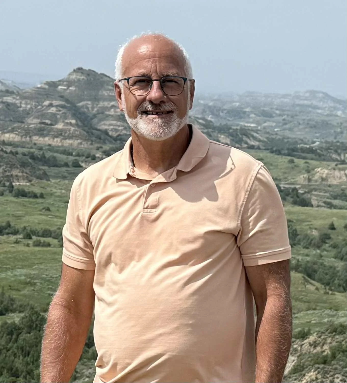 A middle-aged man with glasses, gray hair, and a beard, dressed in a light gray suit, red tie, and a boutonnière, standing outdoors with greenery and wooden structures in the background.