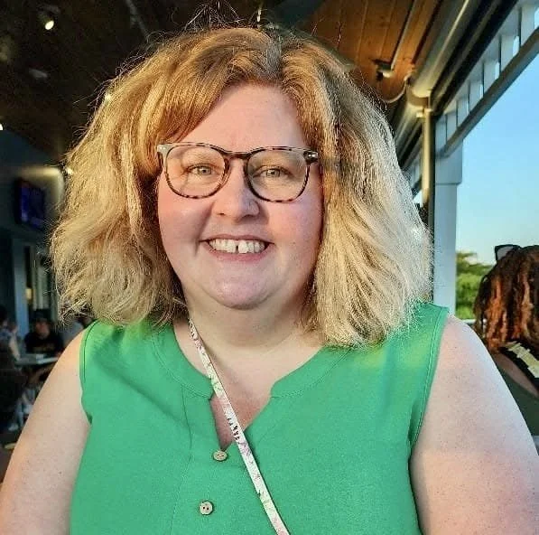 A smiling woman with curly blonde hair and glasses, wearing a green sleeveless top, seated indoors near large windows with natural light.