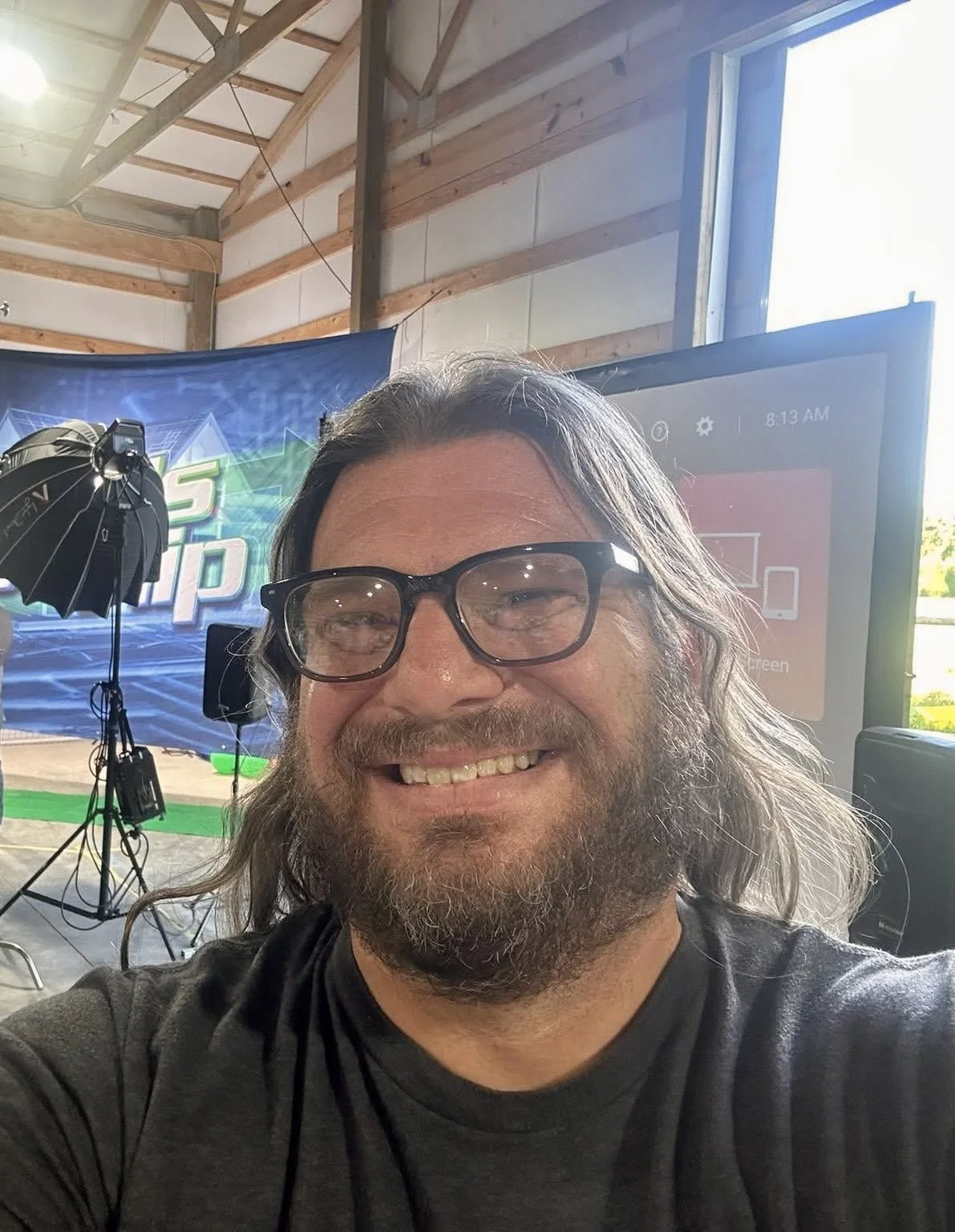 A man with long hair, glasses, and a beard smiling for a selfie in a room with a large monitor and a stage setup in the background.