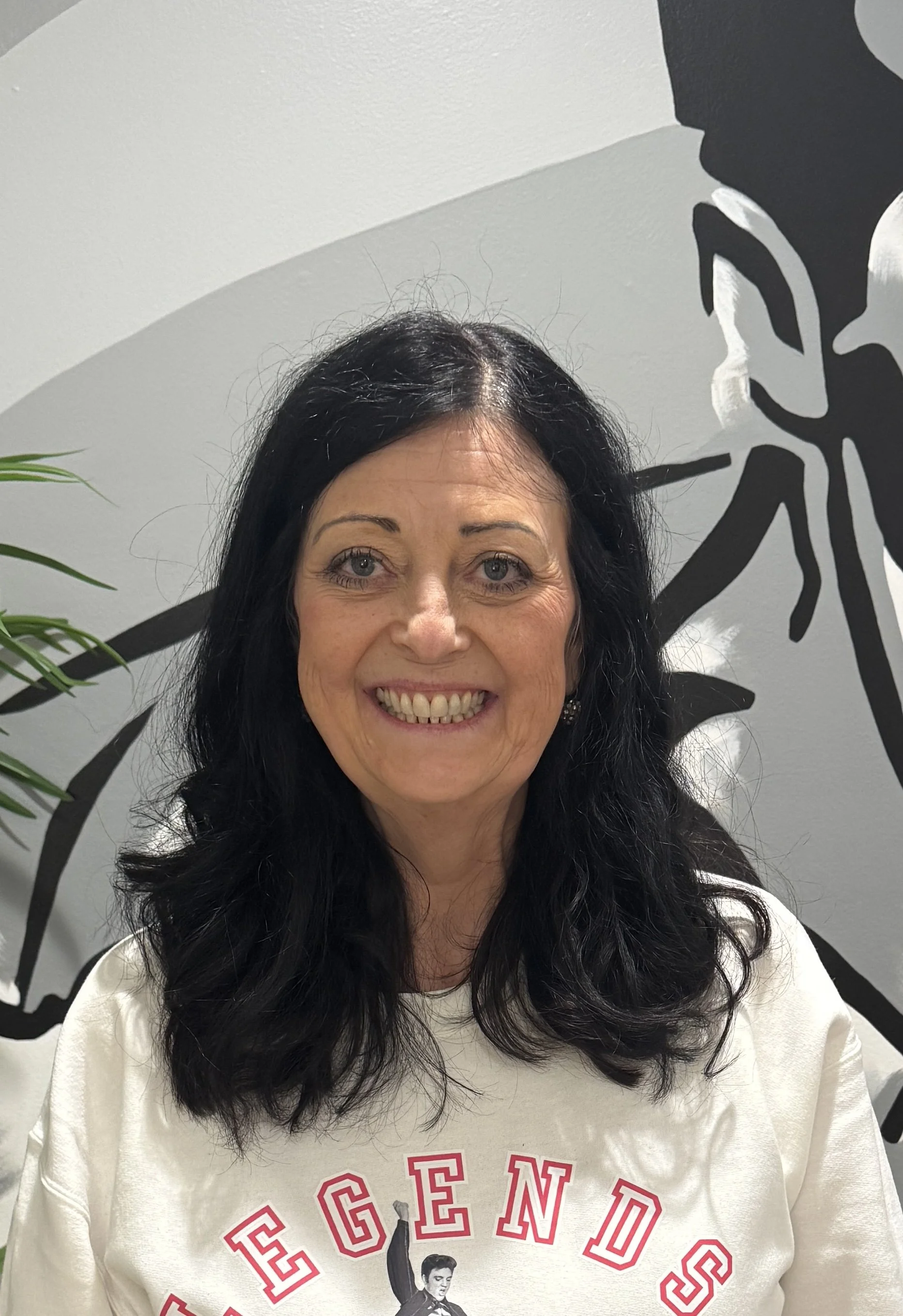 Smiling woman with black hair and a white sweatshirt with red text and an illustration, standing in front of a wall with black and white abstract artwork.