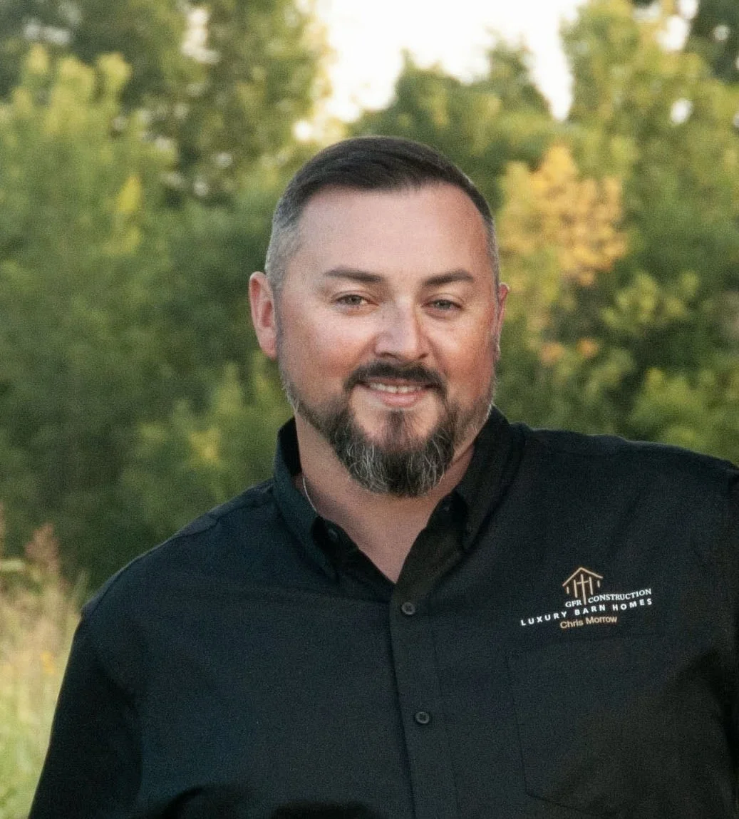 A man with a beard and short hair smiling outdoors, wearing a black shirt with a logo and text that says "GFR Construction Luxury Barn Homes Chris Morrow."