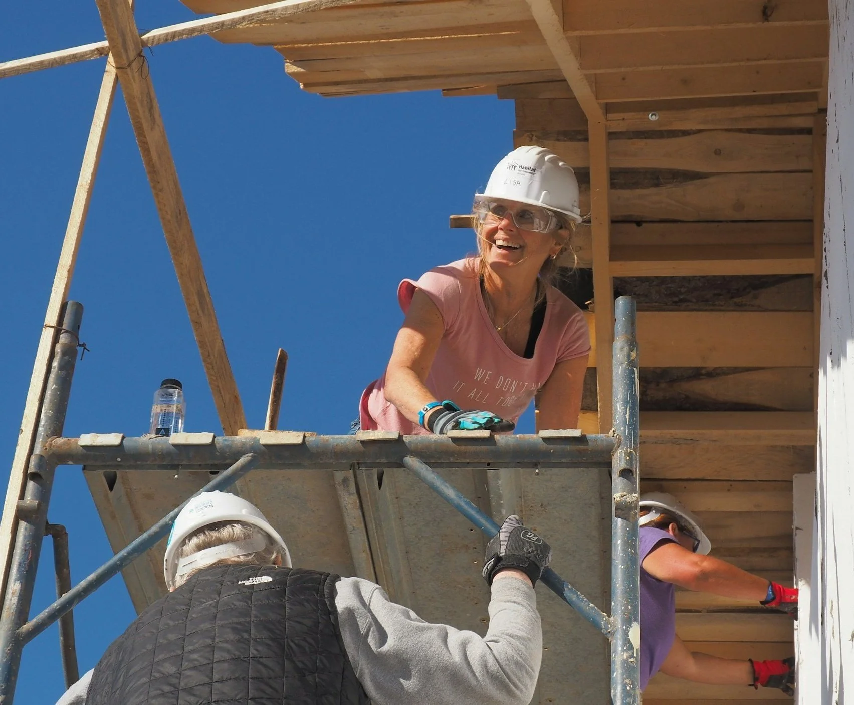 A woman wearing safety glasses and a pink T-shirt, smiling, working on a construction site with scaffolding, under a clear blue sky.