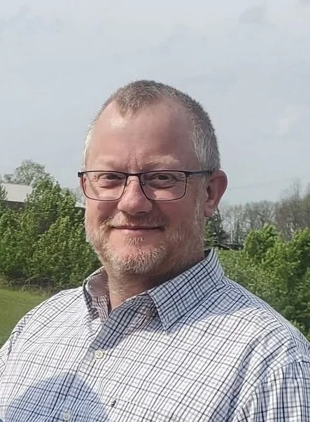 A man with glasses and short gray hair standing outdoors with trees and a cloudy sky in the background.
