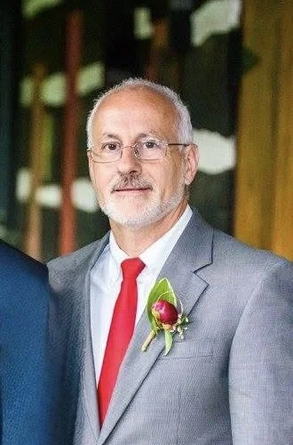 A middle-aged man with glasses, gray hair, and a beard, dressed in a light gray suit, red tie, and a boutonnière, standing outdoors with greenery and wooden structures in the background.