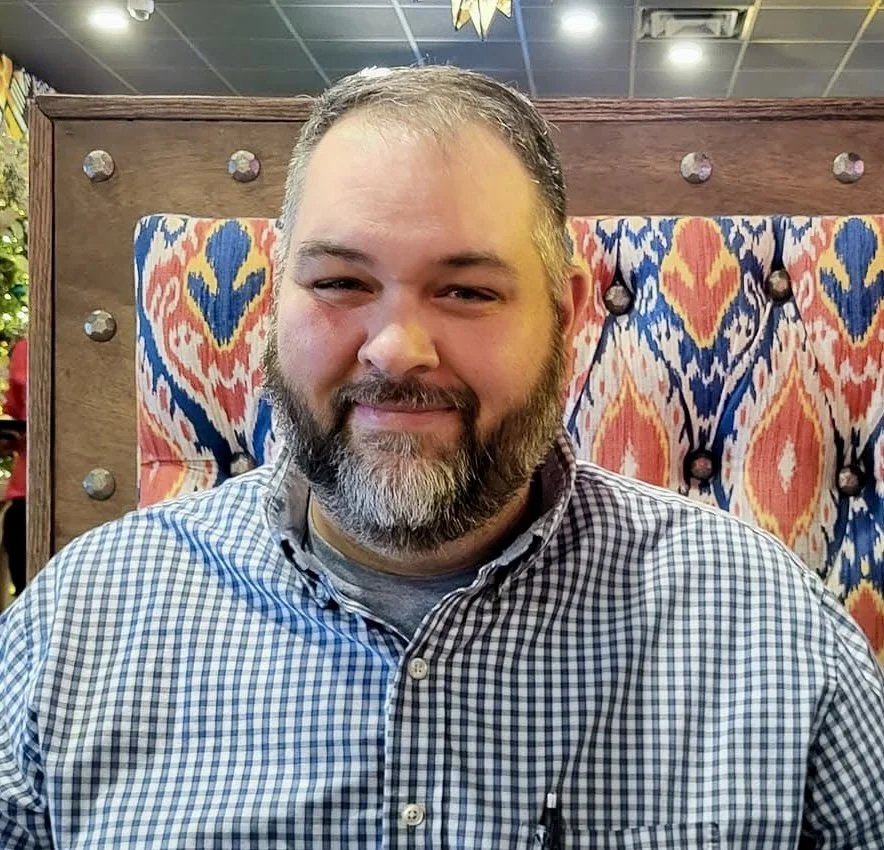 Man with a beard sitting in front of a colorful patterned background, smiling at the camera.